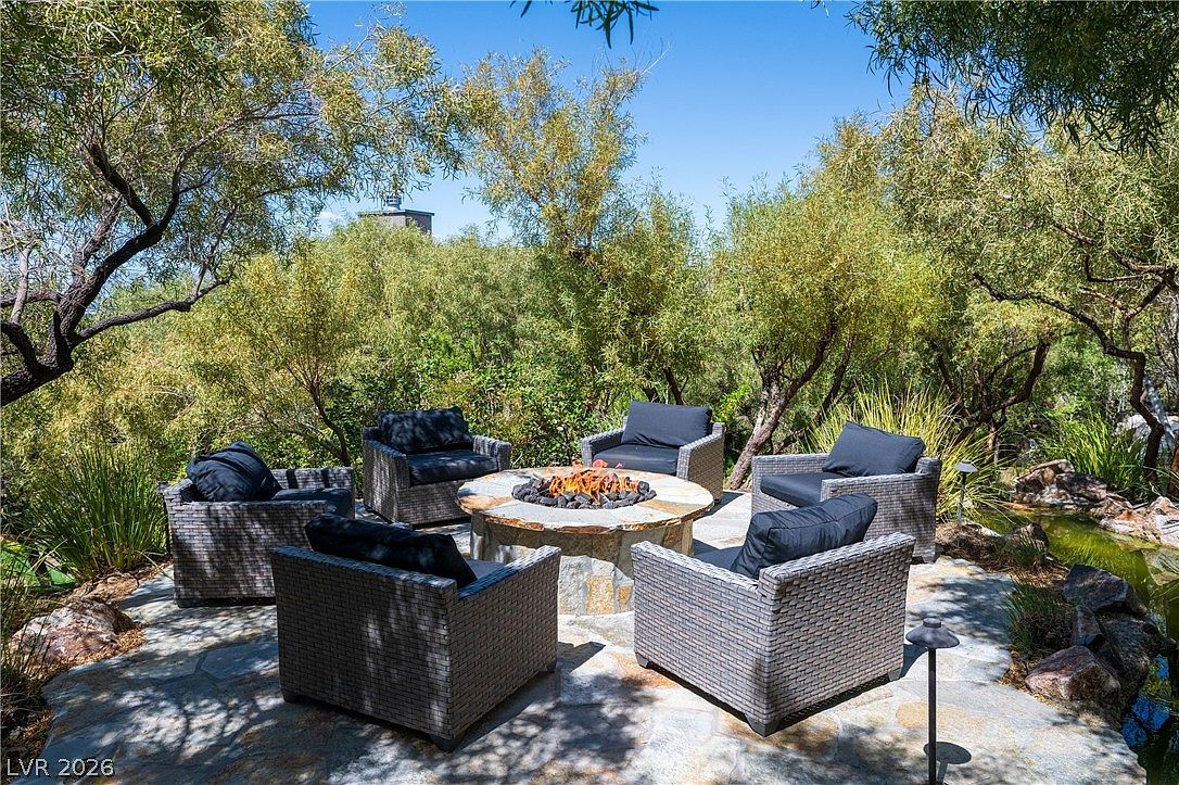 This inviting outdoor patio features a circular stone fire pit as its centerpiece, surrounded by five comfortable wicker-style armchairs with dark cushions. The seating area is set upon a natural stone paver floor, nestled within a lush, tree-filled landscape that provides a sense of privacy and seclusion. A small water feature is visible to the right, enhancing the tranquil atmosphere of this serene backyard retreat.