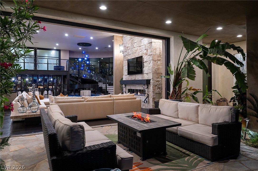 This luxurious covered patio seamlessly blends indoor and outdoor living through a wide opening that reveals a grand interior living space with a piano and dramatic blue lighting. The outdoor area features a central fire table surrounded by plush, dark-wicker seating, set against a backdrop of a tall stone-clad chimney breast and lush tropical greenery. The perspective captures the sophisticated transition between the cozy, fire-lit exterior lounge and the expansive, high-ceilinged interior beyond.