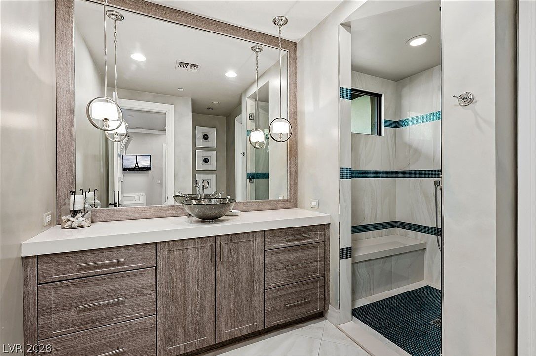 This modern bathroom features a wood-toned vanity with a white countertop and a unique glass vessel sink. A large mirror spans the wall above the vanity, accented by two contemporary pendant lights, while an adjacent walk-in shower includes a built-in bench and decorative blue tile accents. The space is bright and clean, showcasing a sophisticated design with a neutral color palette.