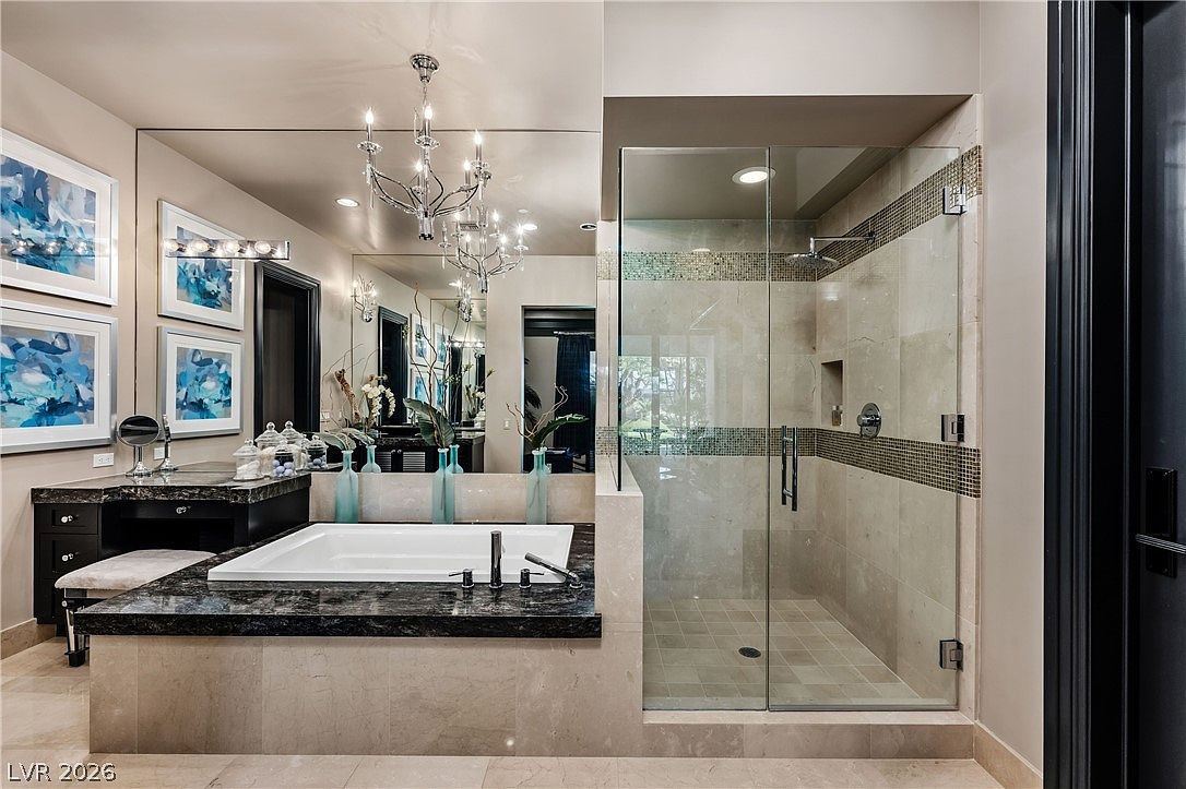 This luxurious primary bathroom features a deep soaking tub set within a dark stone surround, complemented by a glass-enclosed walk-in shower with neutral-toned wall tiles. A large mirror spans the wall above the vanity, reflecting a decorative chandelier and elegant blue glass accents. The space is designed with a sophisticated, high-end aesthetic, emphasizing clean lines and a polished, spa-like atmosphere.