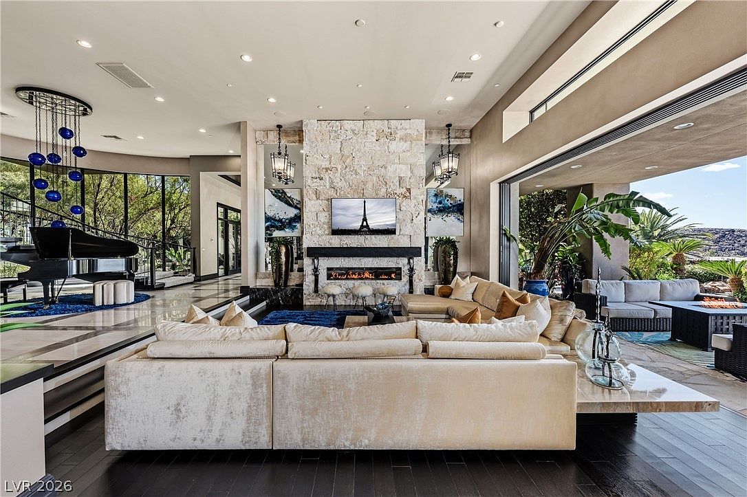 This expansive, open-concept living room features a grand stone-clad chimney breast as its central focal point, flanked by large windows that provide a seamless transition to the outdoor patio. A sleek black grand piano sits on a circular blue rug to the left, while a plush, light-colored sectional sofa anchors the main seating area. The space is defined by high ceilings, recessed lighting, and a sophisticated blend of dark wood flooring and polished stone accents, creating an atmosphere of luxury and refined comfort.