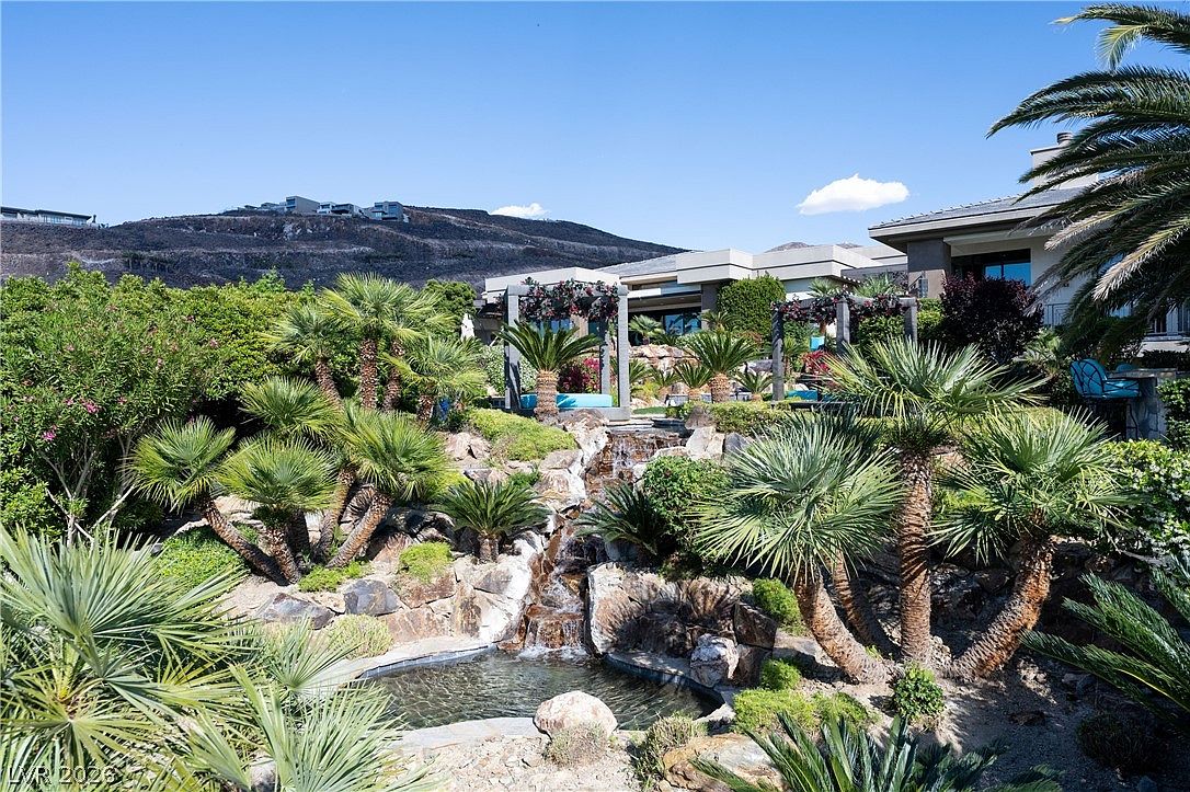 This lush, tiered backyard landscape features a cascading stone waterfall that flows into a small pond, surrounded by mature palm trees and desert-adapted greenery. Two pergola-covered seating areas provide shaded spots for relaxation, while the backdrop showcases a modern residence set against a rugged mountain hillside. The scene captures a serene, resort-style outdoor living space designed for privacy and natural beauty.