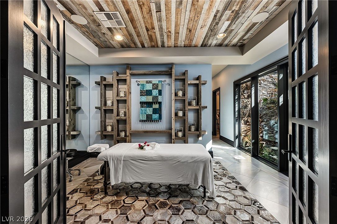 This serene home spa room features a central massage table set upon a distinctive hexagonal-patterned rug. The space is accented by rustic wood-paneled ceilings and symmetrical wall-mounted shelving units flanking a decorative textile wall hanging. Large glass doors provide a view of an outdoor garden area, creating a tranquil and luxurious atmosphere.