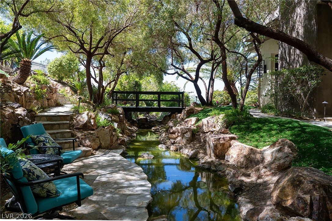 This serene backyard garden features a tranquil stream flowing through a naturalistic landscape of large boulders and mature trees. A wooden footbridge spans the water, connecting the stone-paved patio area, which is furnished with comfortable outdoor seating, to the lush green lawn on the opposite bank. The scene is captured from a low-angle perspective, emphasizing the peaceful, park-like atmosphere of the property.