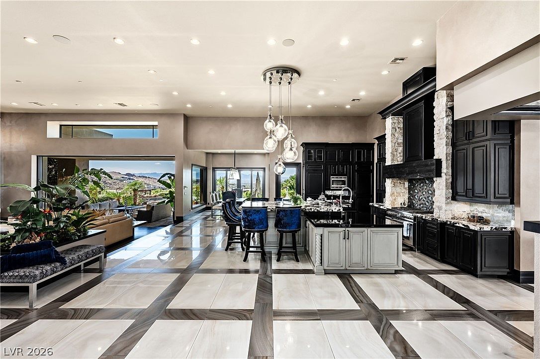 This expansive open-concept kitchen and living area features a striking contrast between dark cabinetry and light, patterned tile flooring. A large central island with blue velvet bar stools serves as the focal point, illuminated by a cluster of modern glass pendant lights. The space flows seamlessly into a living area that opens to a scenic outdoor view through large glass doors, creating a bright and luxurious atmosphere.