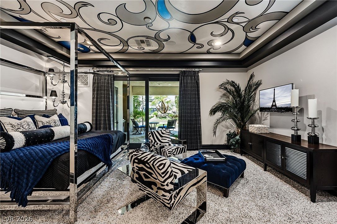 This luxurious primary bedroom features a striking four-poster chrome bed frame and a ceiling adorned with intricate, swirling artistic murals. The room is furnished with two zebra-print armchairs, a plush blue velvet ottoman, and a dark wood media console, all set against a neutral carpet. Large glass sliding doors provide a seamless transition to an outdoor patio, allowing natural light to fill the sophisticated, high-contrast space.
