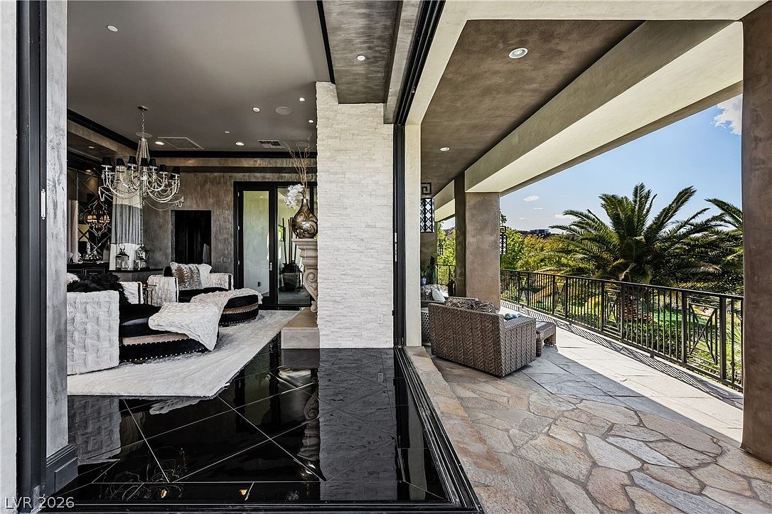 This sophisticated living space features a seamless transition to an outdoor balcony through expansive sliding glass doors. The interior is anchored by a glossy black tiled floor and a prominent stone-clad chimney breast, while the adjacent covered patio offers a stone-tiled floor and a scenic view of palm trees. The overall aesthetic is one of luxury and indoor-outdoor integration, perfect for high-end entertaining.