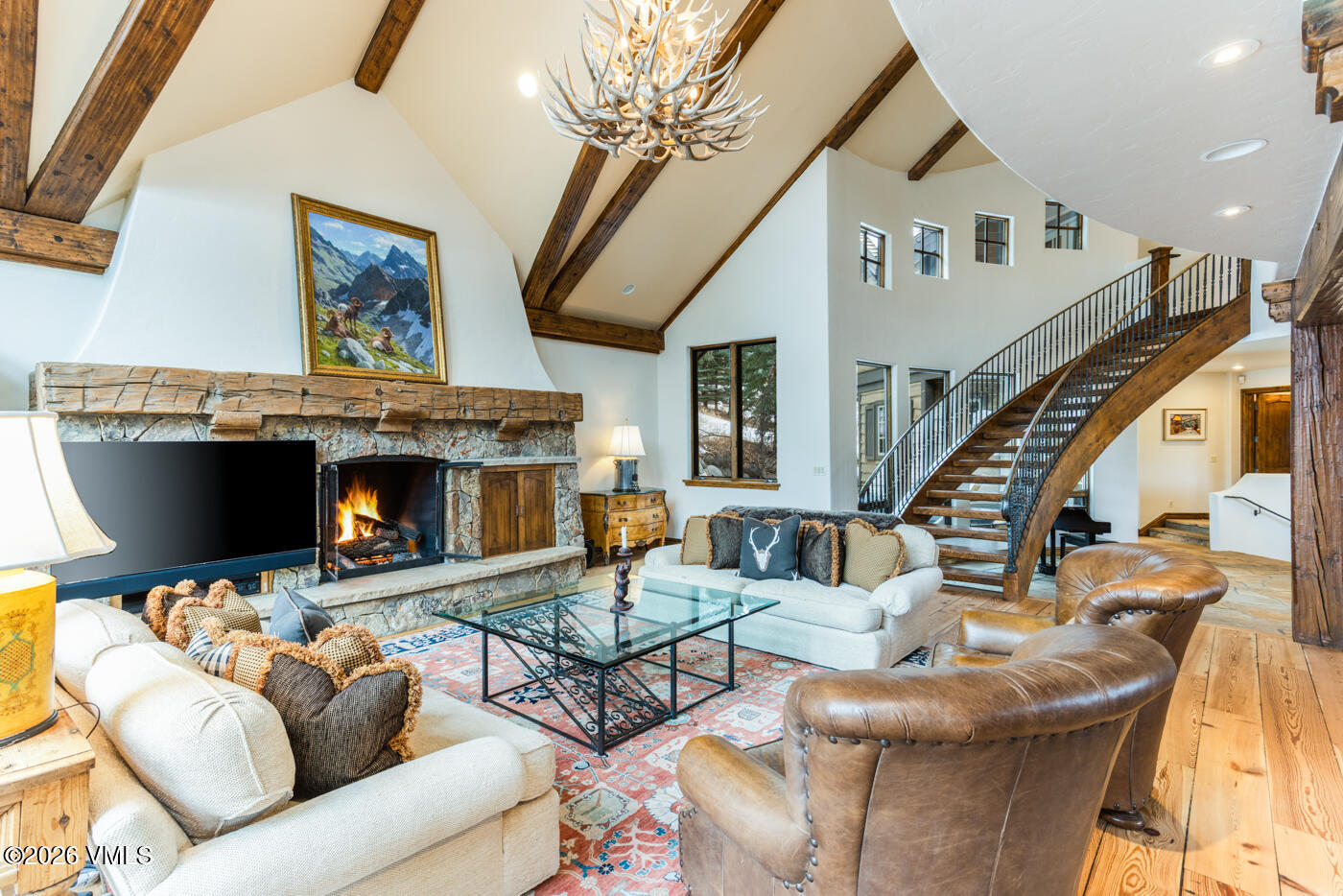 This spacious living room features a vaulted ceiling with exposed wooden beams and a striking antler chandelier. A stone-clad chimney breast serves as the focal point, housing a fireplace and a television, while a grand staircase with a metal railing curves elegantly along the side. The room is furnished with comfortable seating, including two light-colored sofas and two leather armchairs, all arranged on a patterned area rug over hardwood flooring.