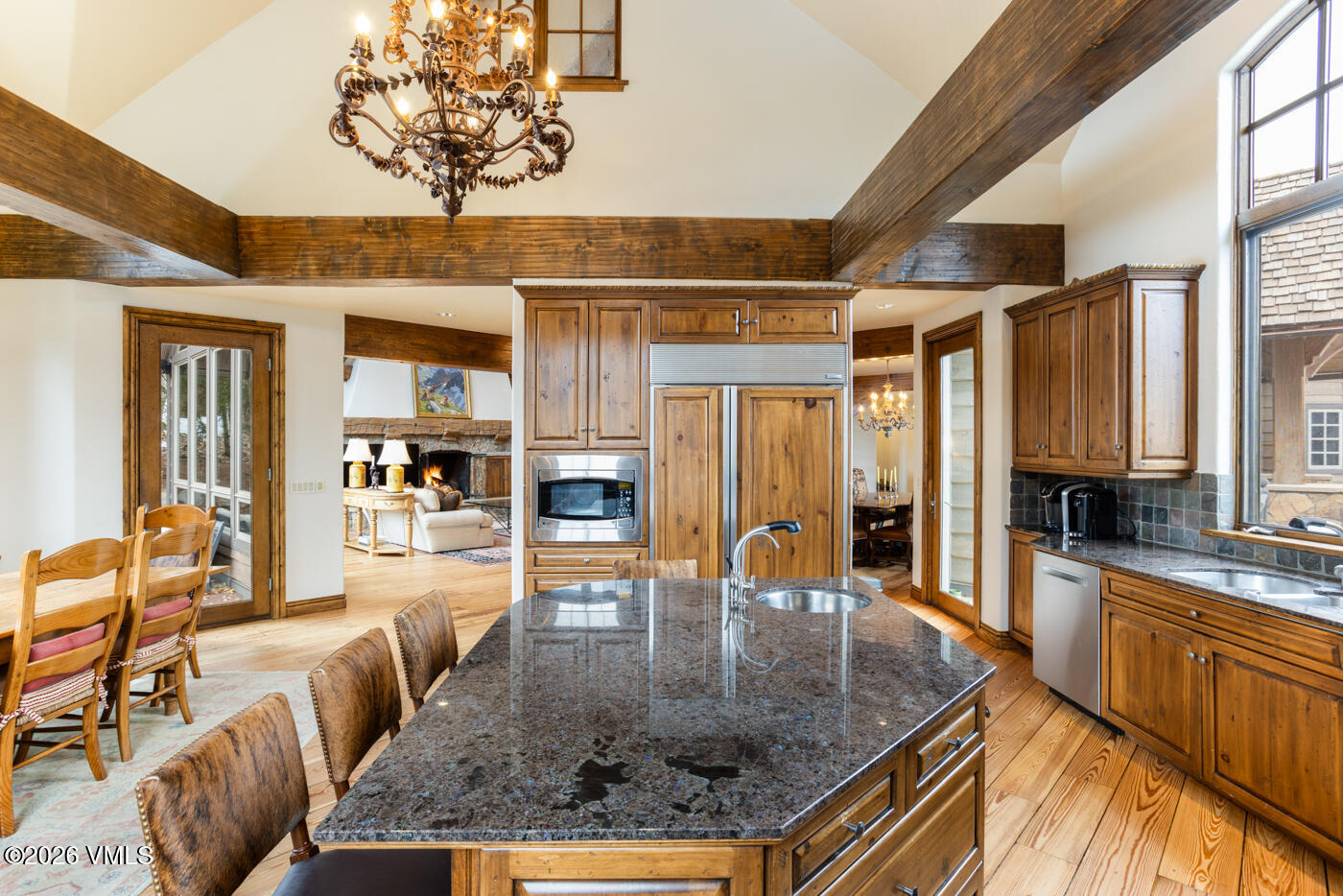 This spacious, rustic-inspired kitchen features warm wood cabinetry, a large central island with a dark stone countertop, and exposed timber ceiling beams. The open-concept layout flows into an adjacent living area with a stone fireplace, creating a cohesive and inviting atmosphere. Large windows provide ample natural light, highlighting the rich textures of the wood and stone throughout the space.