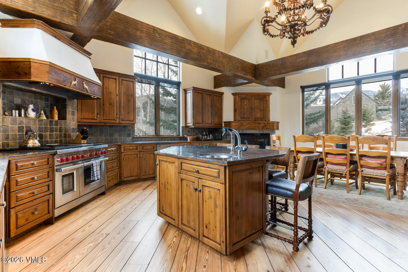 This rustic-style kitchen features warm wood cabinetry, a large central island with a stone countertop, and a professional-grade stainless steel range. The space is defined by exposed timber beams on the vaulted ceiling and wide-plank wood flooring, creating a cozy, mountain-lodge atmosphere. Large windows provide ample natural light and views of the snowy exterior, while a decorative chandelier adds a touch of elegance above the adjacent dining area.