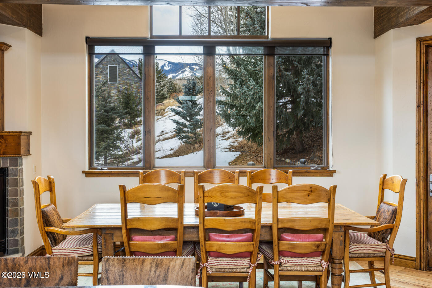 This dining room features a rustic wooden table surrounded by six matching chairs with red-cushioned seats and striped fabric accents. A large, multi-pane window serves as the focal point, offering a scenic view of a snowy, wooded landscape and distant mountains. The room is framed by dark wood ceiling beams and a stone-clad fireplace surround on the left, creating a cozy, mountain-lodge atmosphere.