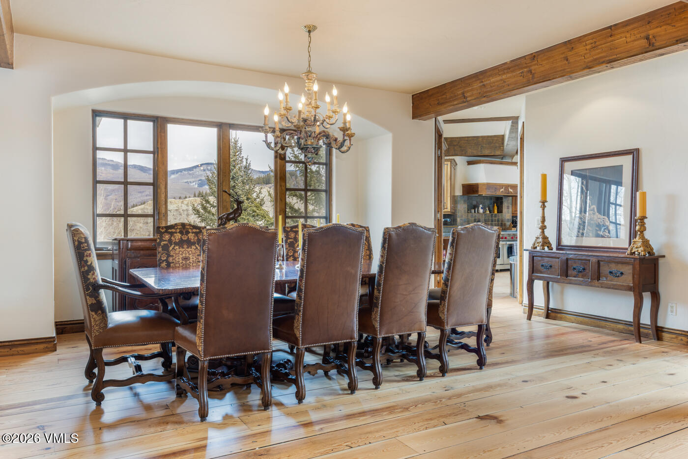 This elegant dining room features a large, dark wood table surrounded by high-backed leather chairs with nailhead trim. A grand, multi-armed chandelier hangs above the table, while a large window set into an arched alcove provides natural light and mountain views. The room is accented by exposed wooden ceiling beams, light wood flooring, and a traditional sideboard topped with decorative lamps and framed artwork.
