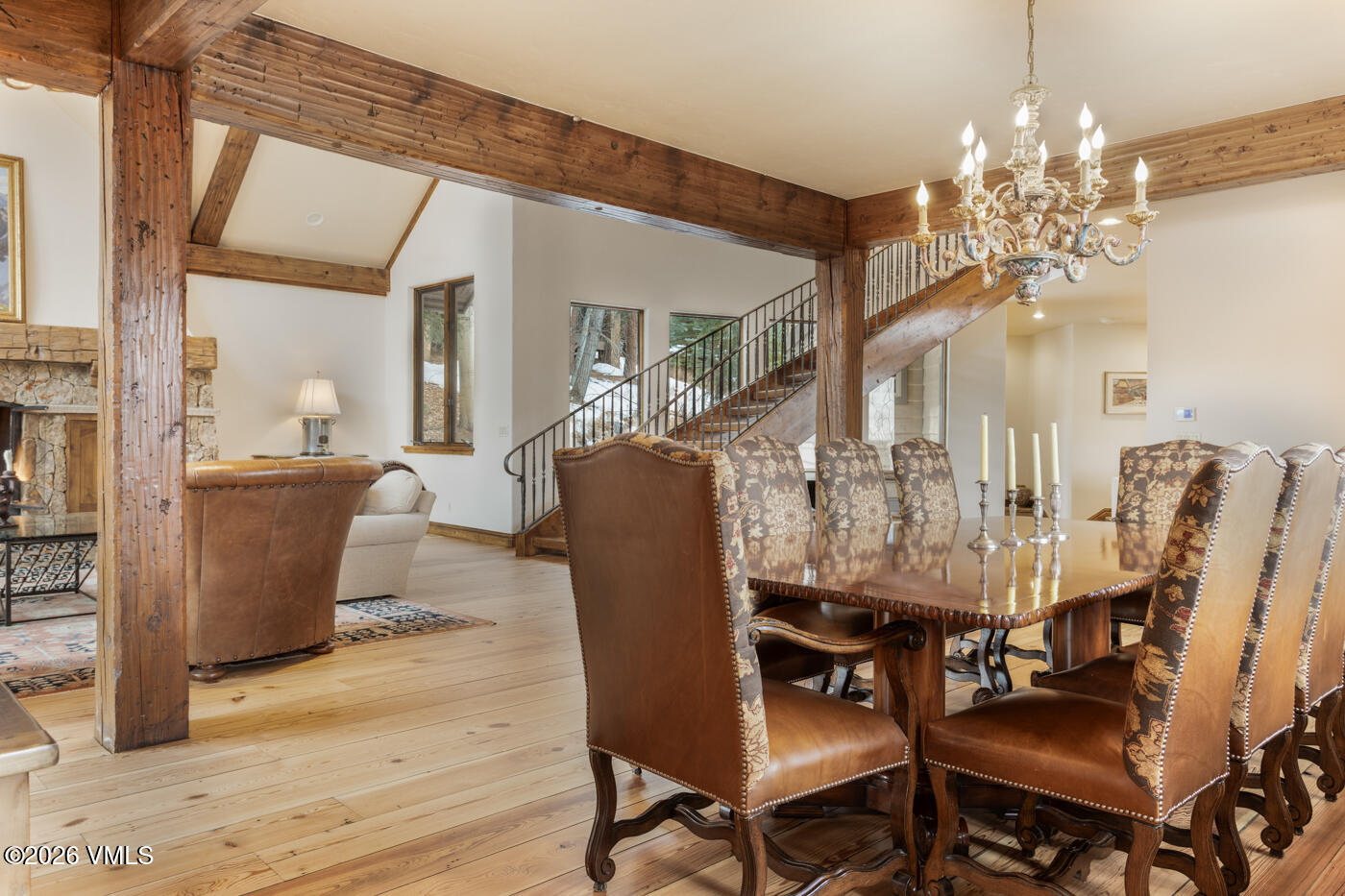 This elegant dining area features a large wooden table surrounded by high-backed chairs with patterned upholstery and leather seats, illuminated by an ornate chandelier. The space is defined by rustic exposed timber beams and light wood flooring, creating a warm, mountain-inspired aesthetic. In the background, a staircase with a metal railing leads to an upper level, while a glimpse of a living area with a stone-clad chimney breast is visible through the open floor plan.