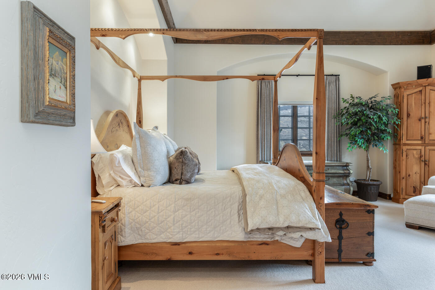 This primary bedroom features a prominent four-poster wooden bed frame as the central focal point, dressed in neutral-toned bedding and plush pillows. The room is accented by rustic wooden ceiling beams, a large potted plant, and a wooden storage chest at the foot of the bed. A window with soft drapery allows natural light to enter, creating a warm and inviting atmosphere.