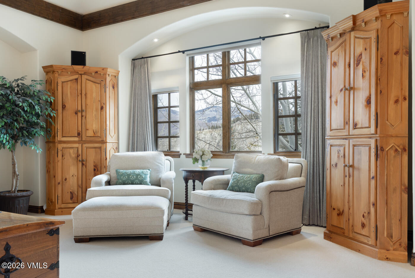 This cozy seating area features two plush, light-colored armchairs, one with an accompanying ottoman, positioned in front of a large, arched window arrangement that offers scenic mountain views. The room is framed by two tall, light wood armoires and accented by exposed wooden ceiling beams, creating a warm and inviting atmosphere. A small, dark wood pedestal table sits between the chairs, topped with a vase of white flowers, enhancing the refined, traditional aesthetic of the space.