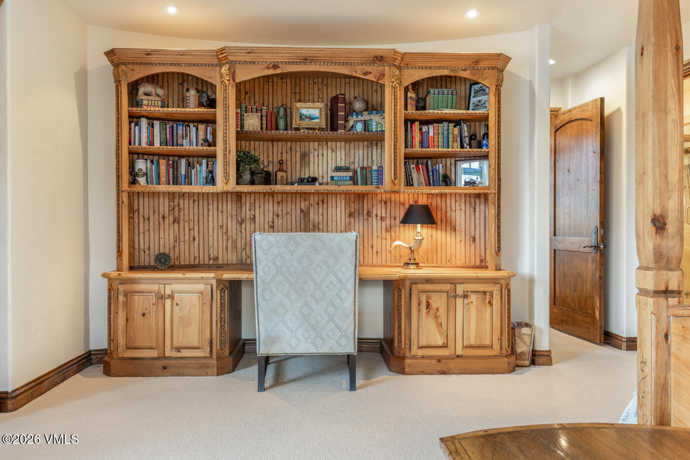 This home office space features a large, custom-built light wood desk unit with integrated shelving and cabinetry. A patterned upholstered chair is positioned at the desk, which is accented by a small lamp and decorative items on the shelves. The room is finished with neutral carpeting and a wooden door leading to an adjacent area, creating a warm and functional workspace.
