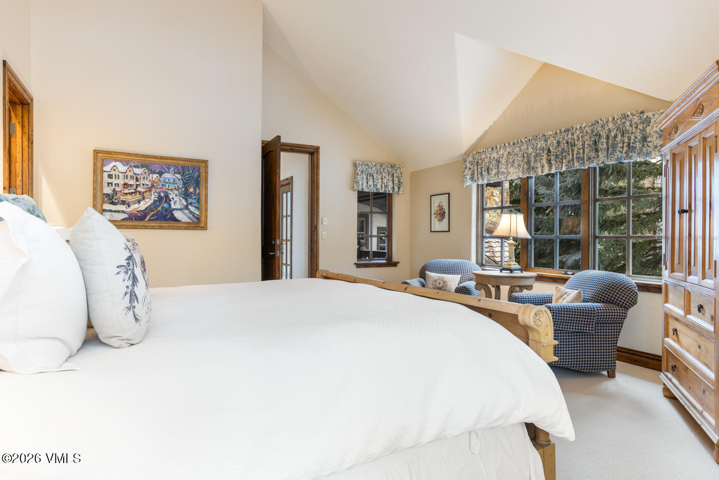 This primary bedroom features a vaulted ceiling and a neutral color palette, creating a bright and airy atmosphere. A large bed with white linens takes center stage, while a cozy seating area with two patterned armchairs and a small round table is positioned near the windows. The room is accented by a wooden armoire, a framed painting, and a valance-topped window arrangement that overlooks the surrounding trees.