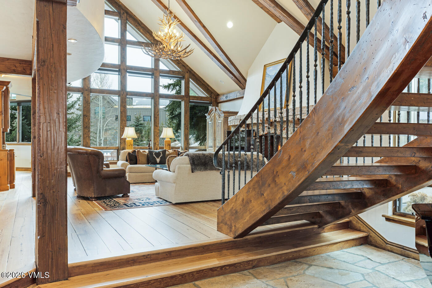 This expansive living room features a rustic mountain aesthetic with exposed timber beams and a vaulted ceiling. A prominent staircase with dark metal railings leads to an upper level, while a large stone-clad chimney breast serves as a focal point near the seating area. The space is bathed in natural light from a wall of large windows that overlook a wooded exterior, creating an airy and inviting atmosphere.