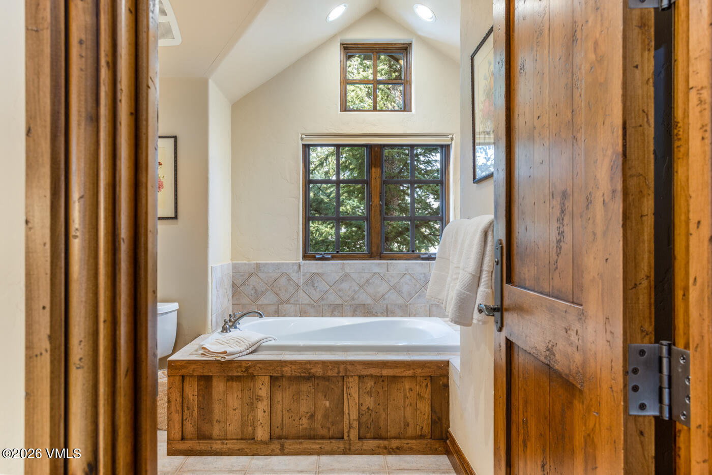 This bathroom features a deep soaking tub encased in rustic wood paneling, set against a backdrop of neutral-toned diamond-patterned tile. The space is illuminated by natural light from a large window and a smaller accent window above, creating a warm and inviting atmosphere. The perspective is framed through a wooden doorway, emphasizing the cozy and private nature of the room.