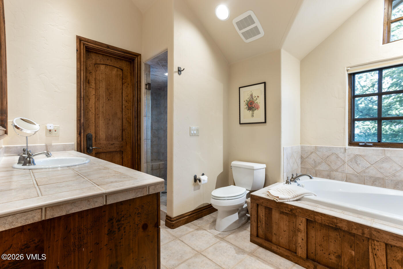 This bathroom features a rustic aesthetic with a wood-paneled vanity and matching bathtub surround. The space includes a white porcelain sink, a separate toilet, and a walk-in shower visible through a doorway, all set against neutral-toned walls and tiled flooring. A window with a grid pattern allows natural light to brighten the room, which is accented by a framed botanical print on the wall.