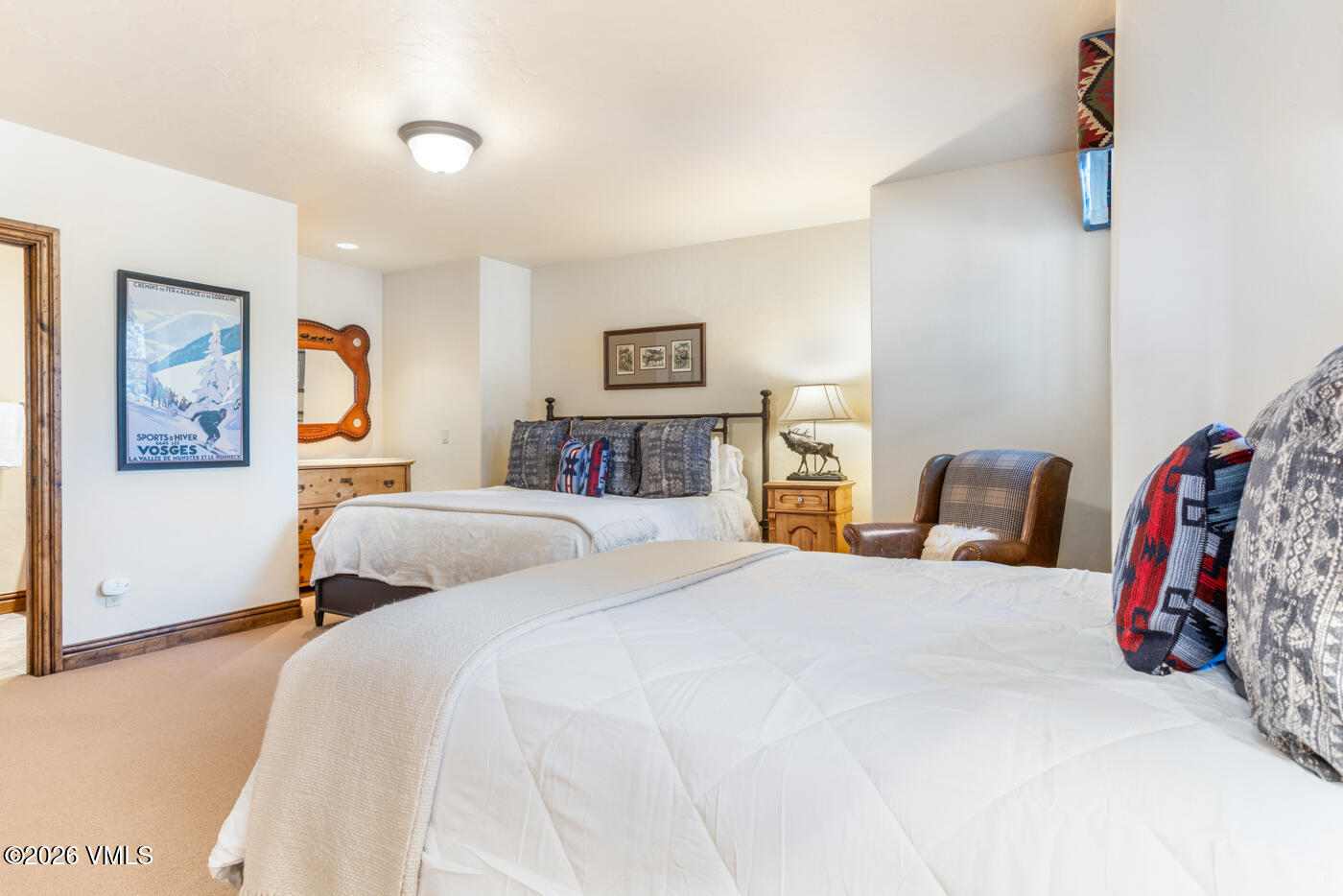 This spacious bedroom features two queen-sized beds dressed in neutral white bedding with patterned accent pillows, creating a cozy and inviting atmosphere. The room is decorated with rustic-inspired elements, including a framed vintage-style ski poster, a wooden dresser with a mirror, and a leather armchair. The warm lighting from the bedside lamp and overhead fixture enhances the comfortable, lodge-style aesthetic of the space.