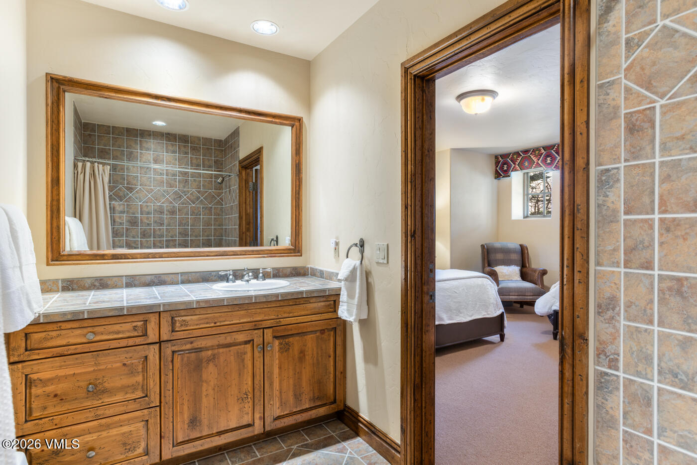 This bathroom features a rustic vanity with a stone-topped counter and a large, wood-framed mirror. The space transitions directly into an adjacent bedroom, which is visible through the open doorway and contains a bed and a comfortable armchair. The overall aesthetic is warm and traditional, with earthy tones and textured tile work.