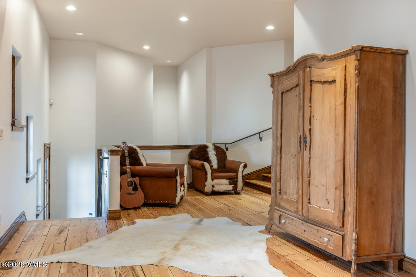This rustic-style landing area features warm wood flooring and a large, light-colored hide rug. The space is furnished with two leather armchairs accented with fur, a wooden wardrobe, and an acoustic guitar leaning against the stair railing. The perspective captures a cozy, inviting transition space that leads toward a staircase, illuminated by recessed ceiling lighting.