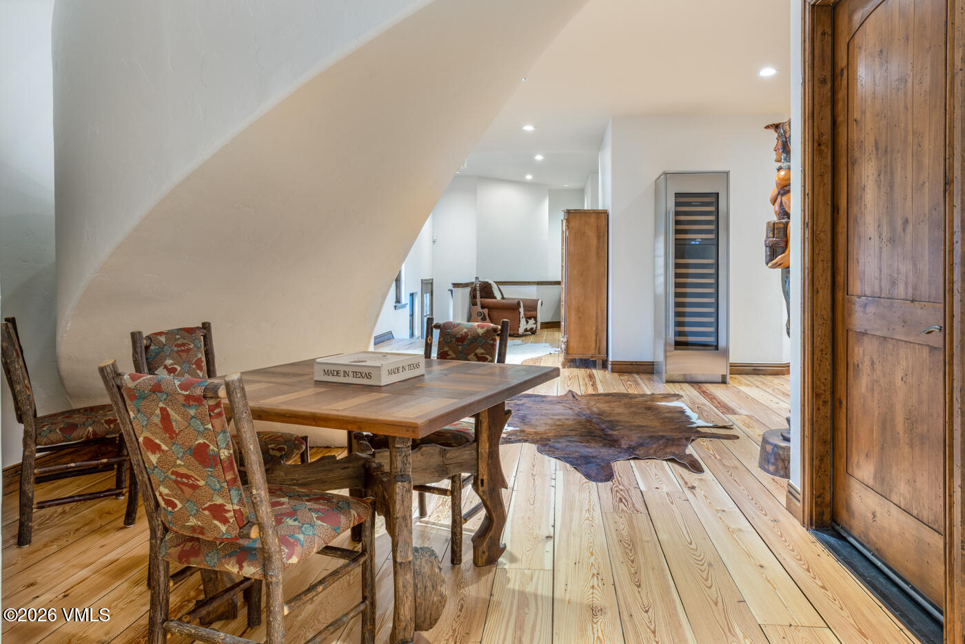 This rustic dining area features a wooden table surrounded by chairs with patterned upholstery and branch-style frames, set against a curved white wall. The space includes light wood flooring, a cowhide rug, and a tall wine refrigerator, leading into an adjacent living area with a leather armchair. The overall aesthetic is warm and western-inspired, with a unique architectural curve defining the room's transition.