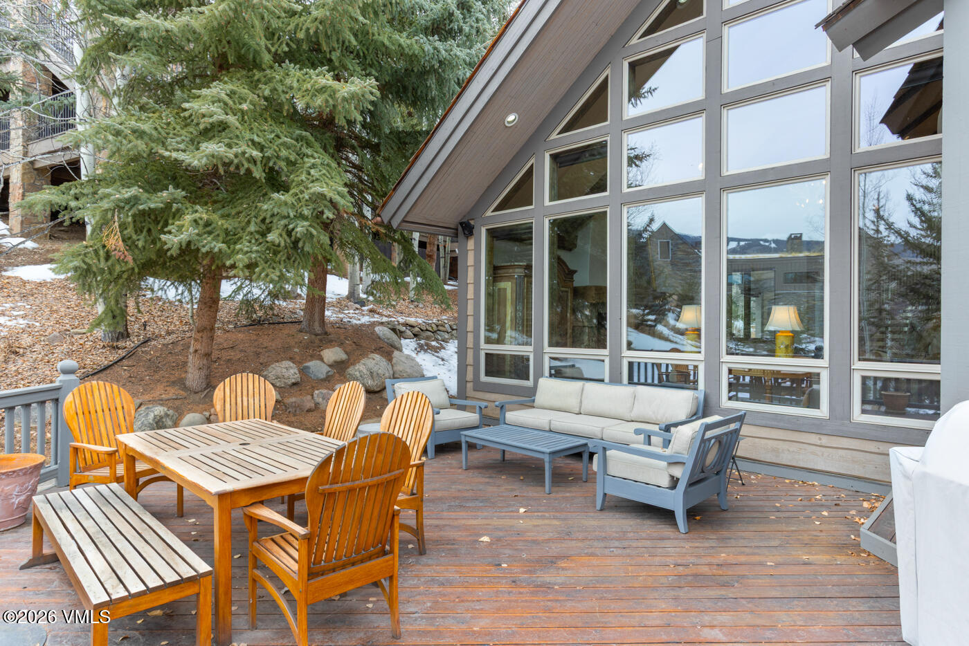 This spacious wooden deck features a dining set with a bench and chairs, alongside a comfortable outdoor seating area with a sofa and armchairs. The deck is set against a backdrop of mature evergreen trees and a sloped, rocky landscape, with large windows providing a view into the home's interior. The overall atmosphere is serene and perfect for outdoor relaxation in a mountain-style setting.
