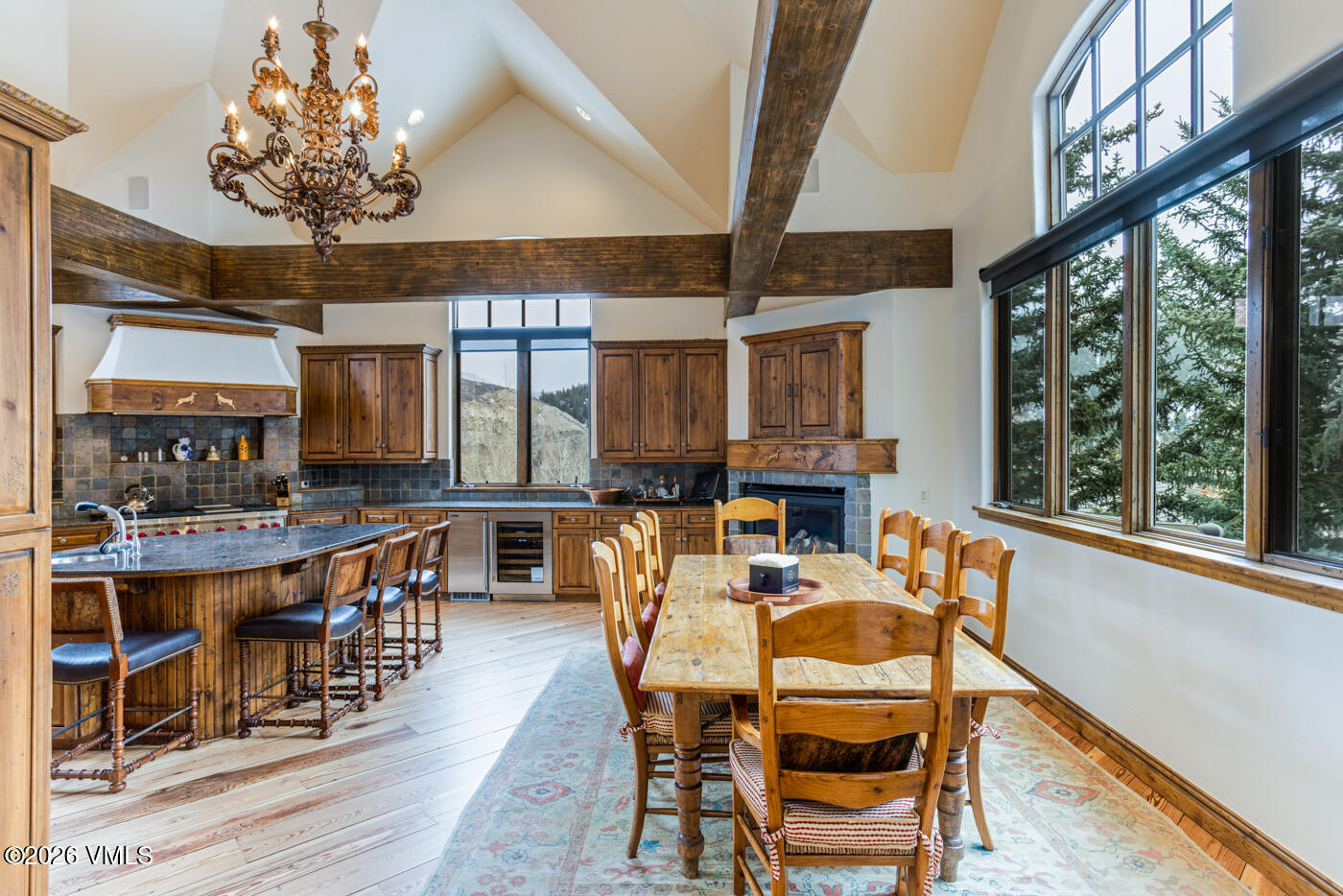 This spacious, open-concept kitchen and dining area features a rustic mountain aesthetic with exposed timber beams and warm wood cabinetry. A large island with a dark stone countertop provides ample seating, while a dedicated dining table sits nearby on a patterned area rug. The room is bathed in natural light from large windows that frame the outdoor scenery, complemented by a decorative chandelier and a stone-clad fireplace.