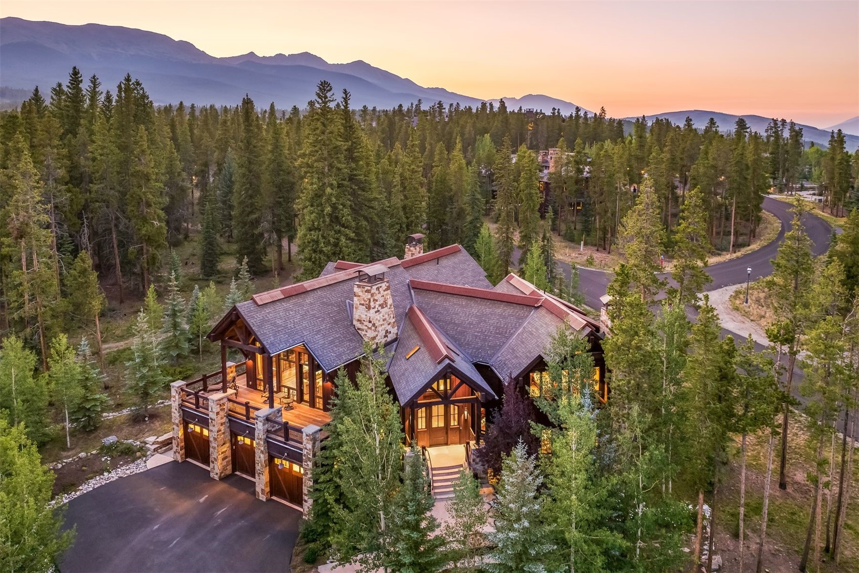 This high-angle aerial perspective captures a luxurious mountain lodge nestled within a dense evergreen forest at sunset. The home features a complex roofline with dark shingles, a prominent stone-clad chimney, and a spacious elevated deck overlooking the driveway and garage area. The surrounding landscape is lush with pine and aspen trees, set against a backdrop of distant mountain peaks under a soft, golden-hour sky.