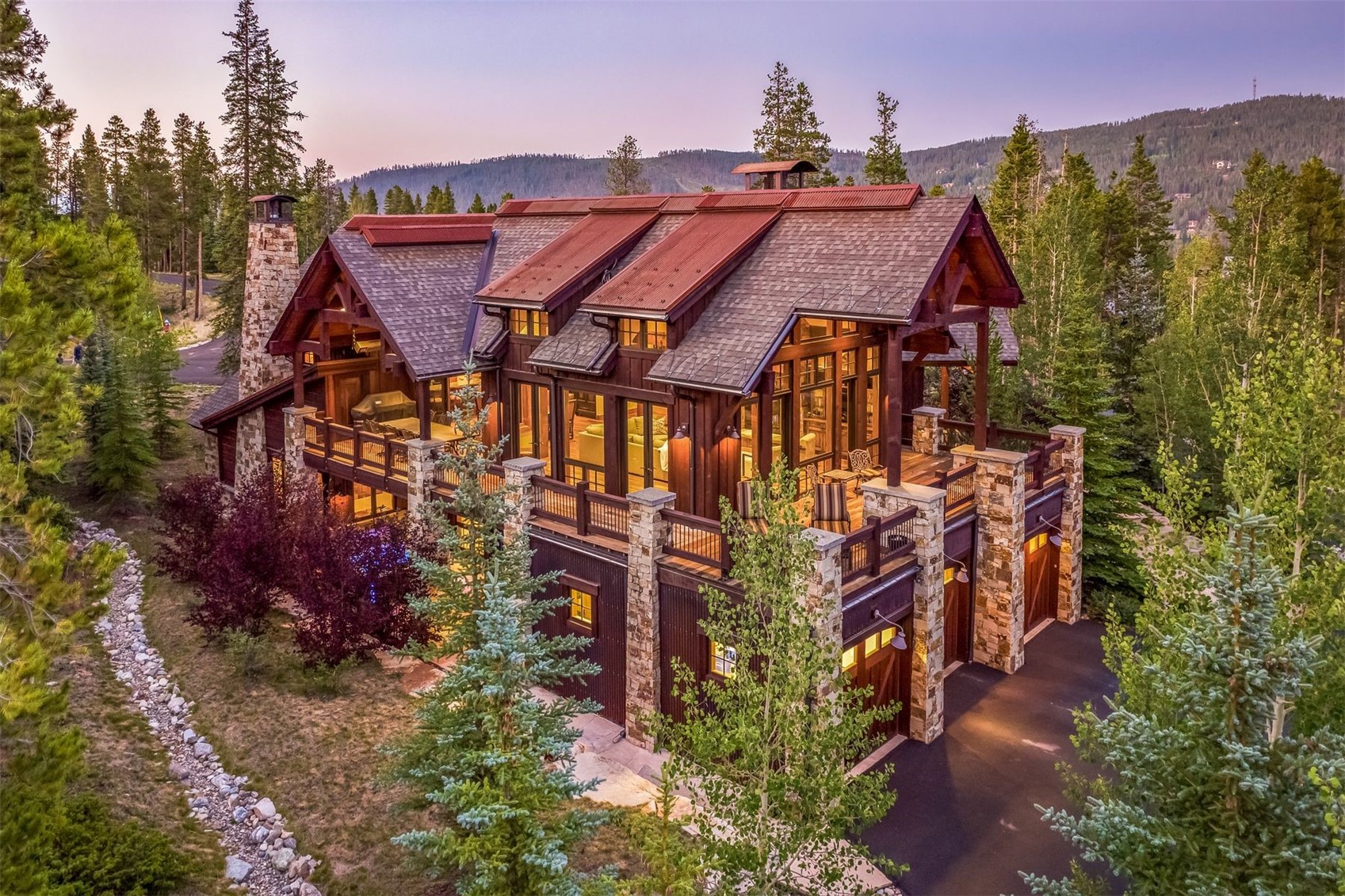 This elevated perspective captures a luxurious mountain lodge nestled within a dense forest at dusk. The home features a complex roofline with metal accents, extensive wood siding, and a prominent stone chimney, complemented by a spacious wrap-around deck and integrated garage bays. The warm interior lighting glows through large windows, creating an inviting atmosphere against the backdrop of the twilight sky.