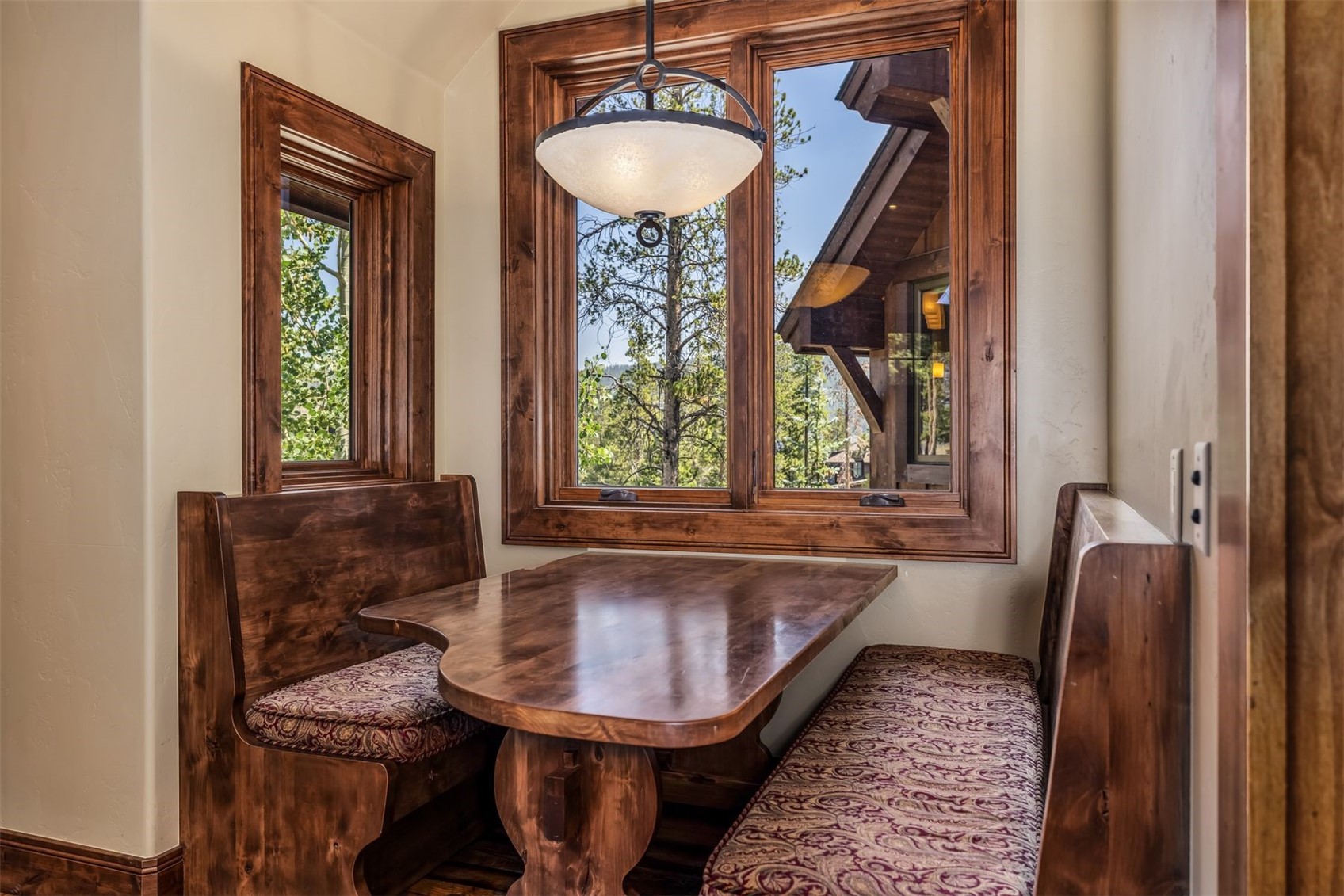 This cozy breakfast nook features a custom-built wooden booth with patterned upholstered cushions, centered around a matching wooden table. A decorative pendant light hangs above the table, while large windows with rich wood trim provide a scenic view of the surrounding trees and exterior architecture. The space is warm and inviting, showcasing a rustic, cabin-inspired aesthetic.