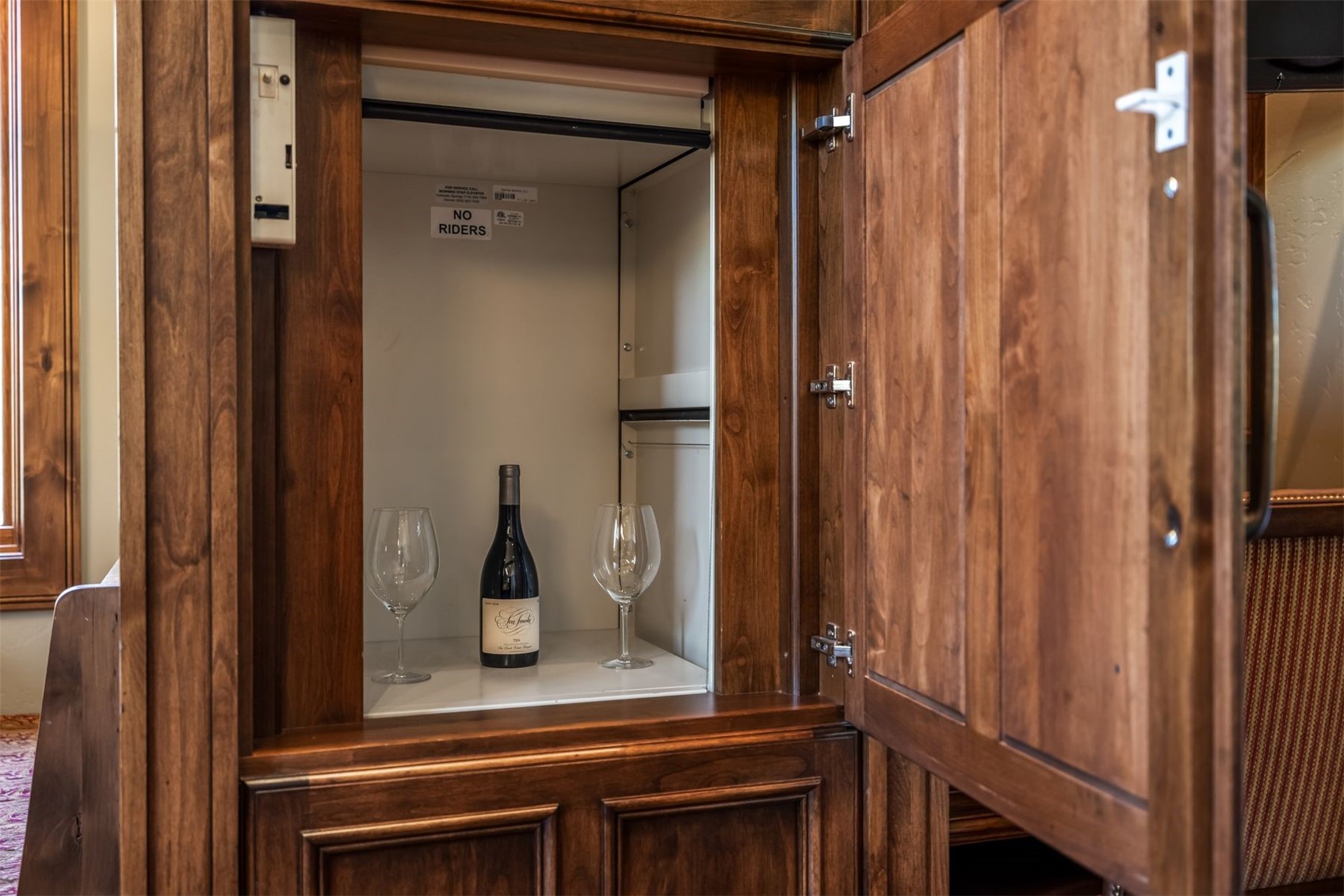 This image captures a custom-built wooden cabinet that houses a small residential dumbwaiter or service lift. The interior of the lift is finished in a clean, neutral material and currently holds a bottle of wine flanked by two empty wine glasses. The rich, dark wood cabinetry features traditional paneling and high-quality hardware, blending seamlessly into the surrounding interior architecture.