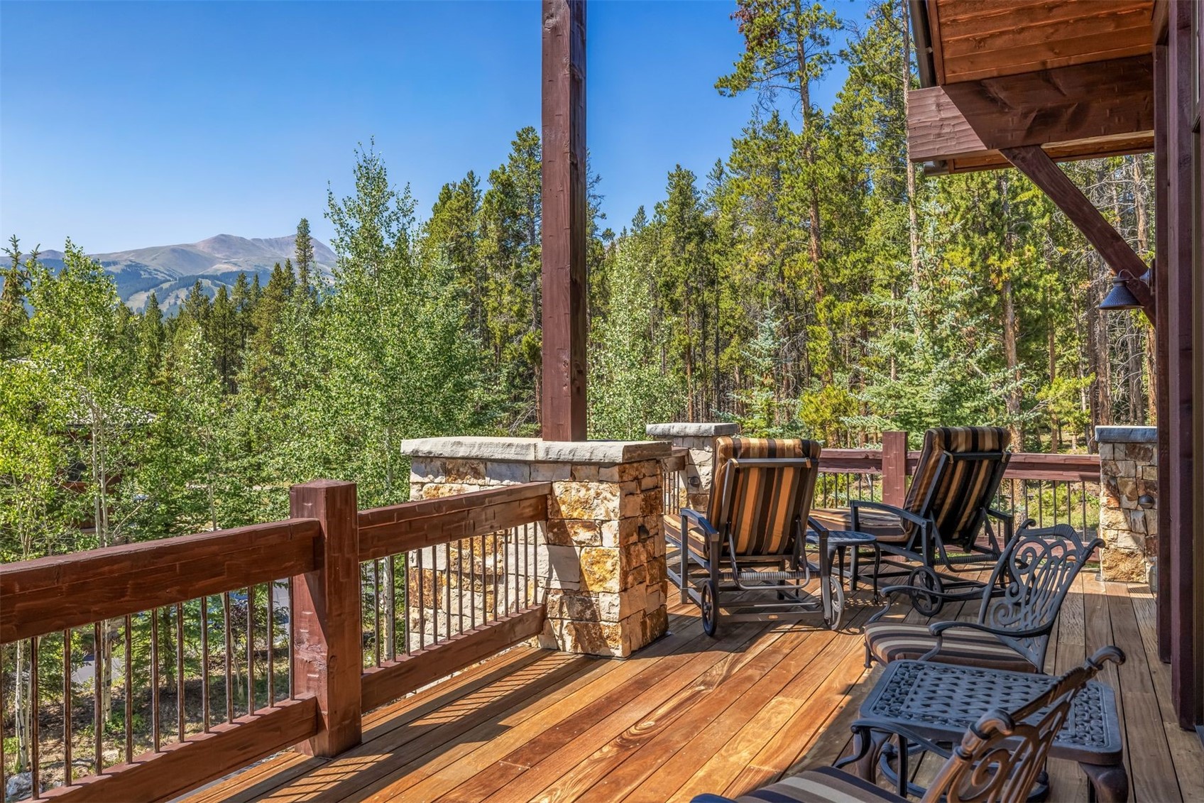 This elevated wooden deck offers a serene outdoor living space with a rustic aesthetic, featuring a sturdy railing with metal balusters and stone-clad support pillars. Several comfortable lounge chairs with striped cushions are arranged to take advantage of the expansive, scenic view of the surrounding pine forest and distant mountains. The perspective is from the deck surface, looking outward to emphasize the connection between the home and the natural landscape.