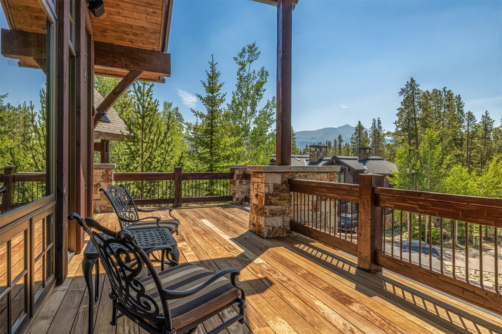 This elevated wooden deck offers a serene outdoor living space with a rustic, mountain-inspired aesthetic. The area is furnished with two metal-framed chairs featuring striped cushions, positioned to overlook a lush forest and distant mountain peaks. Sturdy stone-clad pillars support the covered roof structure, while a wooden railing with metal balusters provides safety and clear sightlines to the natural surroundings below.