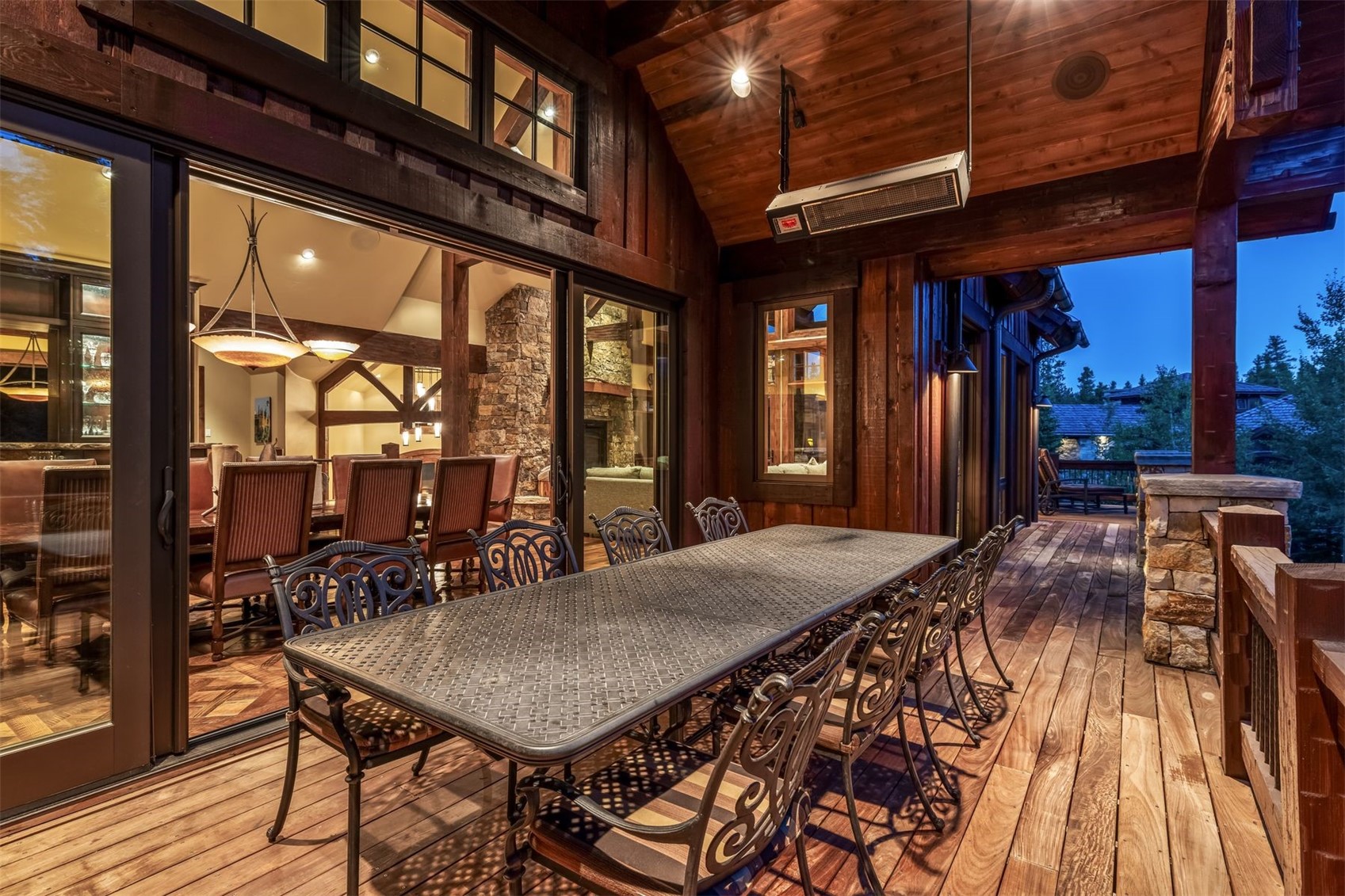 This rustic outdoor deck features a large metal dining set, perfect for al fresco entertaining under a covered, wood-paneled ceiling. Large sliding glass doors provide a seamless transition to the interior dining room, while a stone-clad chimney breast is visible through the glass. The warm wood tones and ambient lighting create an inviting, cozy atmosphere for evening relaxation.