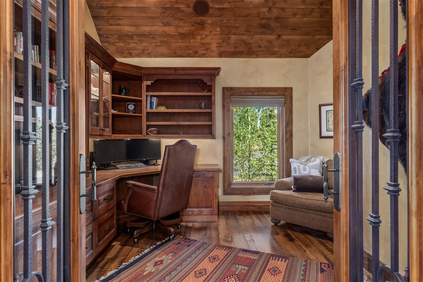 This cozy, rustic-style home office is viewed through a set of decorative metal-framed glass doors, creating a sense of depth and privacy. The room features warm wood-paneled ceilings, rich wood cabinetry with built-in shelving, and a leather desk chair positioned at a corner workstation. A patterned area rug sits on the hardwood floor, while a comfortable armchair near the window provides a quiet space for reading with a view of the outdoors.