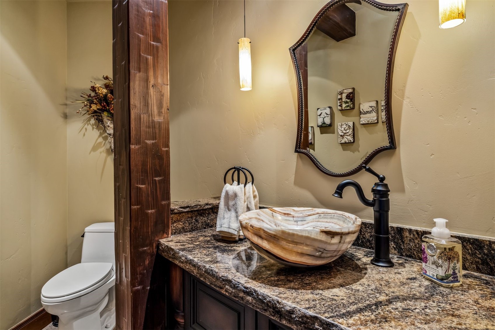 This rustic-style powder room features a unique stone vessel sink set atop a dark granite countertop. A prominent, textured wooden support beam adds character to the space, which is further accented by a decorative mirror and warm pendant lighting. The room includes a white toilet and is decorated with small framed art pieces, creating a cozy and inviting atmosphere.
