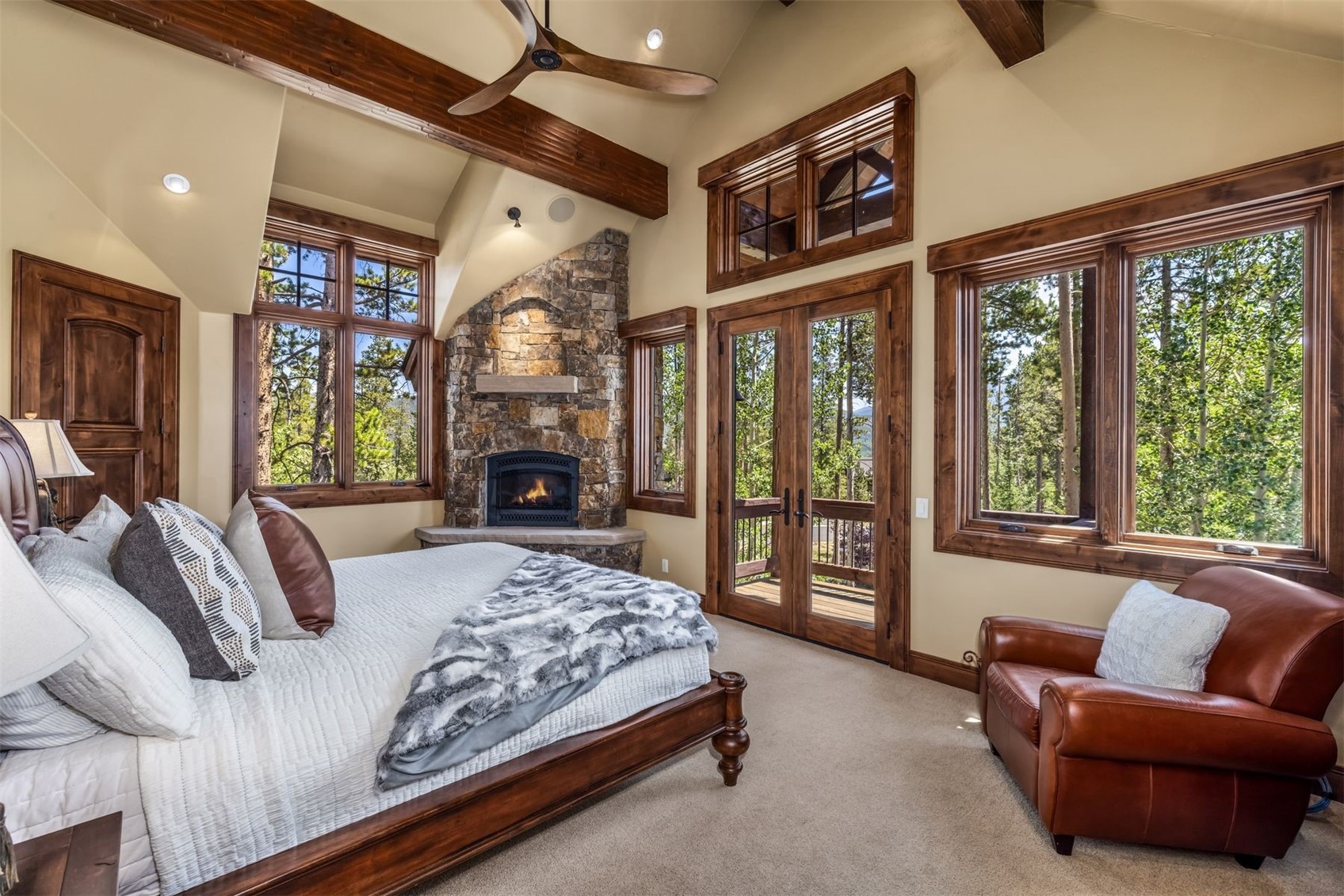 This primary bedroom features a rustic, mountain-inspired aesthetic with a prominent stone-clad chimney breast housing a fireplace. The room is furnished with a large bed topped with a textured throw and a comfortable leather armchair positioned near tall windows that offer views of the surrounding forest. Exposed wooden beams on the vaulted ceiling and rich wood trim around the windows and doors enhance the warm, cozy atmosphere.