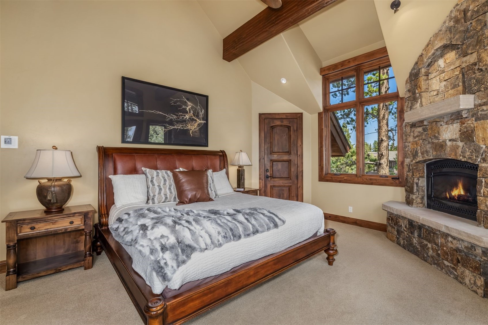 This primary bedroom features a rustic, lodge-inspired aesthetic with a prominent wood-framed bed and a stone-clad chimney breast housing a fireplace. The room is accented by exposed wooden ceiling beams, warm-toned walls, and large windows that provide a view of the surrounding trees. A plush, patterned throw blanket rests on the bed, contributing to the cozy and inviting atmosphere of the space.