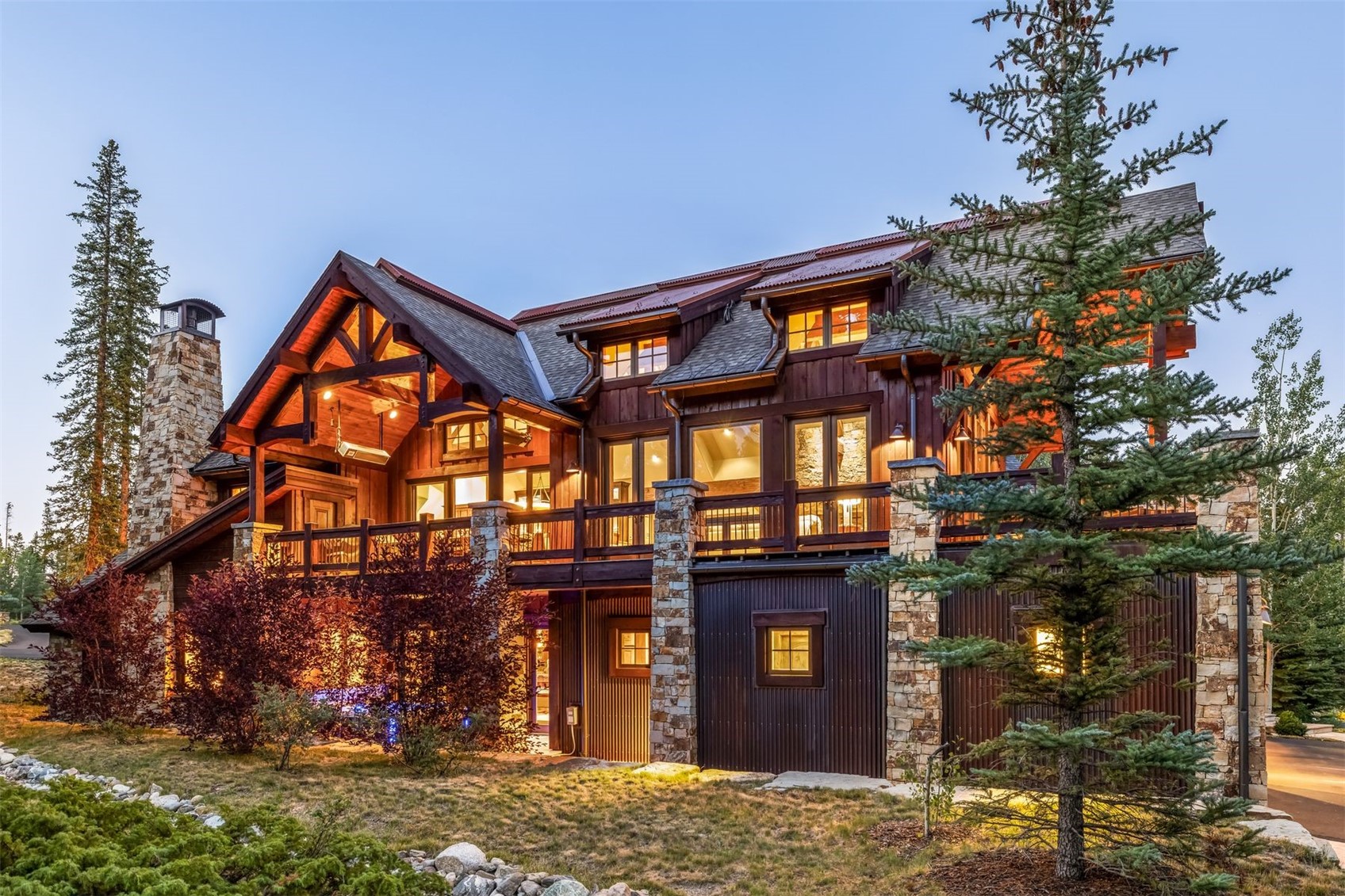 This impressive mountain-style residence showcases a rustic aesthetic with a combination of dark wood siding, corrugated metal accents, and extensive stone masonry. The multi-level structure features a prominent covered deck, a tall stone chimney breast, and numerous windows that glow warmly against the twilight sky. A large evergreen tree stands in the foreground, framing the home's architectural details and expansive outdoor living spaces.