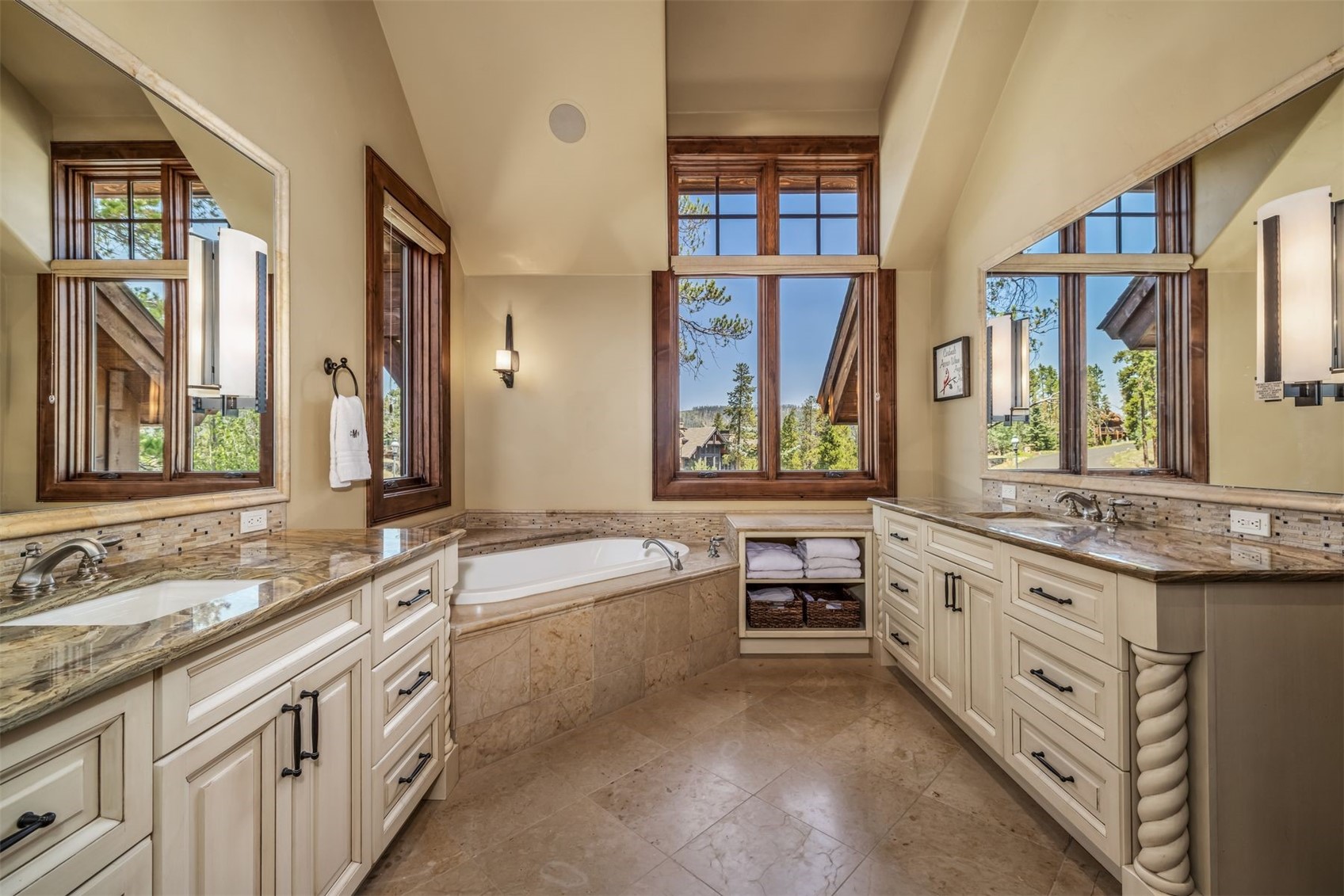 This elegant primary bathroom features dual vanities with light-toned cabinetry and patterned stone countertops, flanking a central corner soaking tub. Large windows with wood trim provide abundant natural light and scenic outdoor views, while the space is finished with neutral stone tile flooring. The room offers a sophisticated, spa-like atmosphere with symmetrical lighting fixtures and integrated storage shelving.