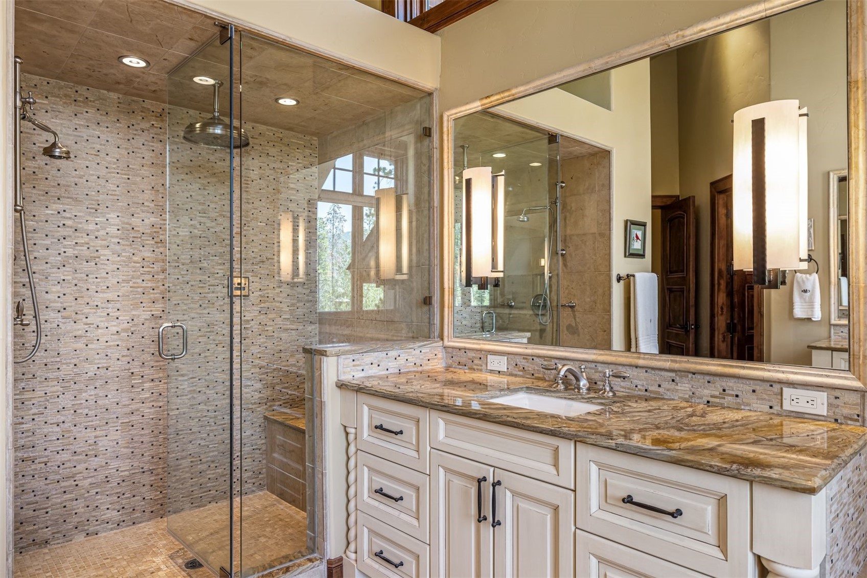 This elegant primary bathroom features a spacious glass-enclosed shower with intricate mosaic tile walls and a built-in bench. A large vanity with a patterned stone countertop and dual wall-mounted light fixtures sits beneath a wide mirror, creating a bright and sophisticated atmosphere. The warm color palette and high-end finishes provide a luxurious, spa-like feel to the space.