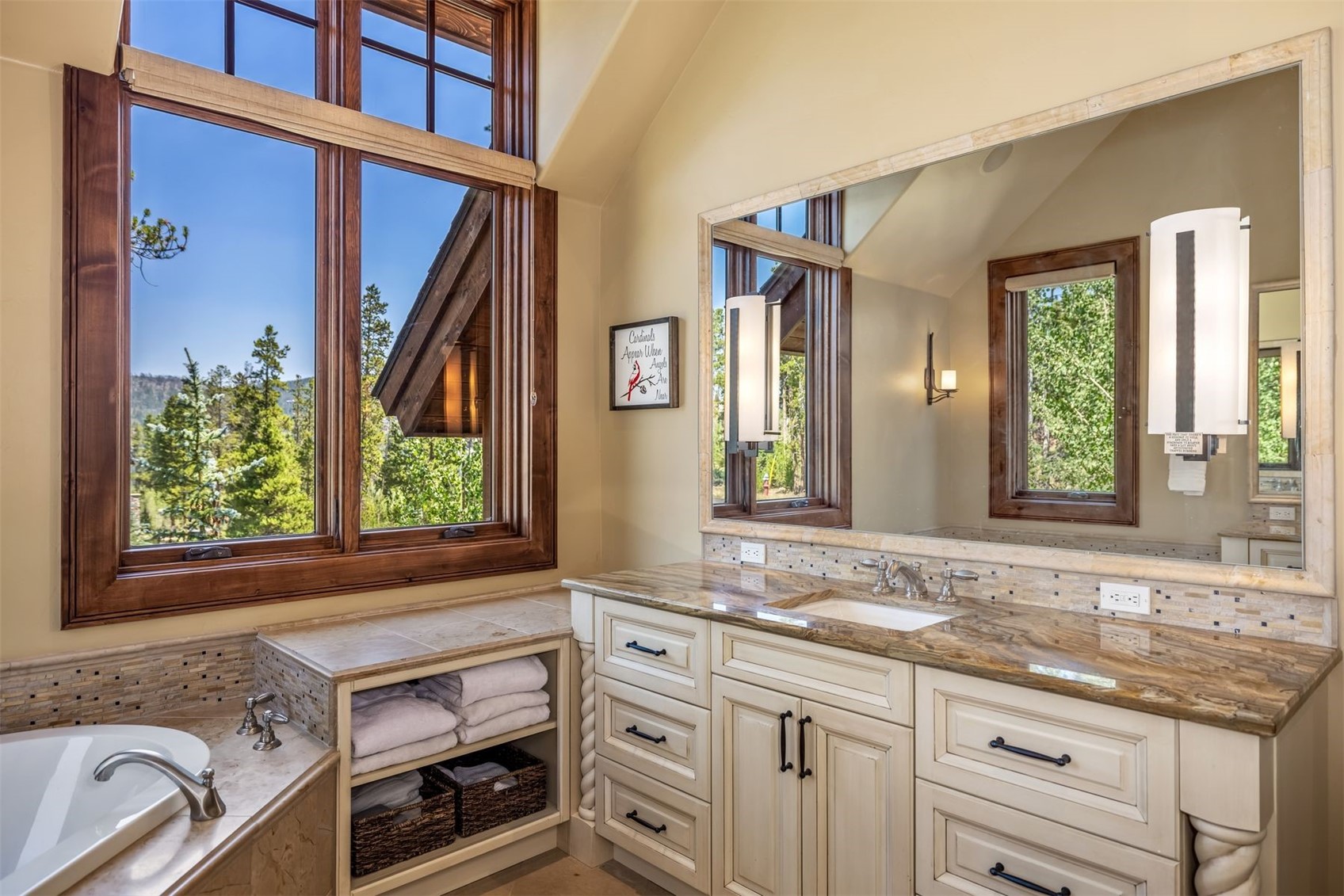 This primary bathroom features a warm, inviting aesthetic with light-toned cabinetry and a patterned stone countertop. A large mirror reflects the tall windows that provide ample natural light and views of the surrounding trees, while built-in shelving keeps towels and baskets neatly organized. The space is accented by elegant light fixtures and a framed wall art piece, creating a serene and well-appointed atmosphere.