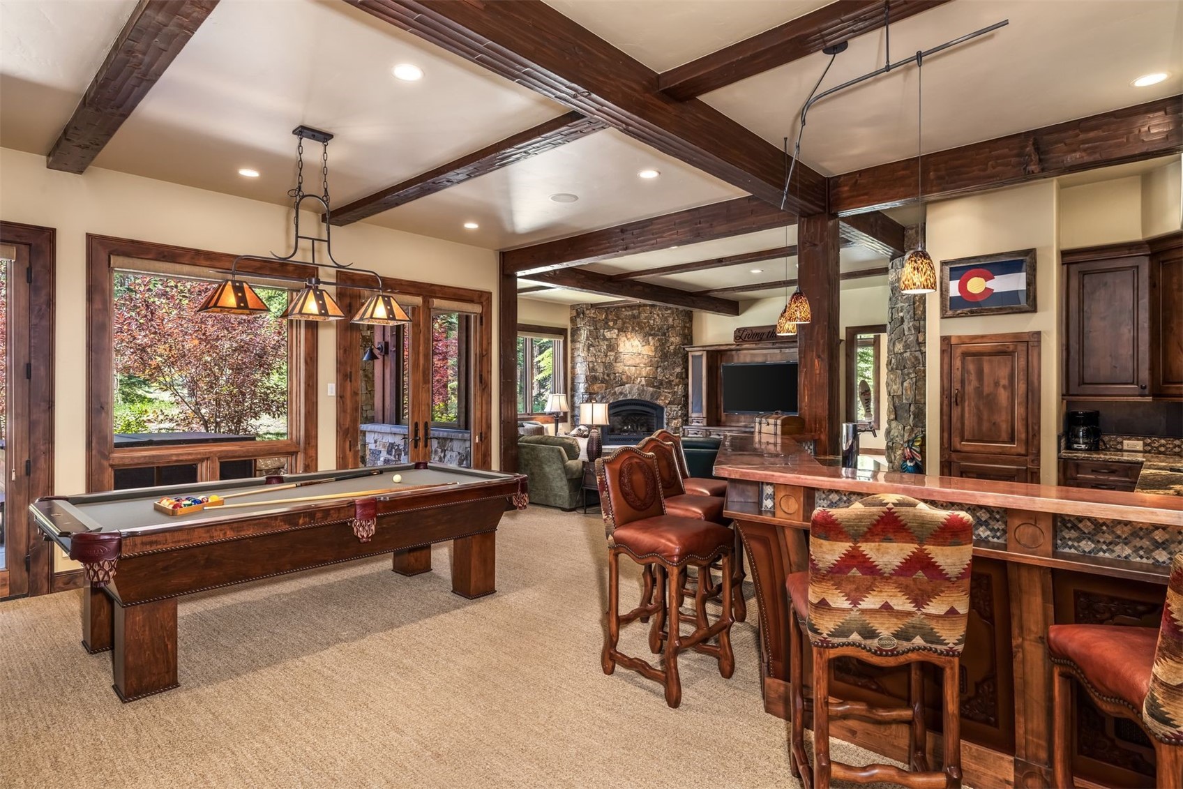 This spacious basement recreation area features a pool table, a custom wood bar with leather-upholstered stools, and a cozy living area with a stone-clad chimney breast. The room is accented by dark wood ceiling beams, warm lighting, and a Colorado flag displayed on the wall, creating a rustic and inviting atmosphere. Large windows provide a view of the outdoors, while the open layout seamlessly connects the gaming and lounge spaces.