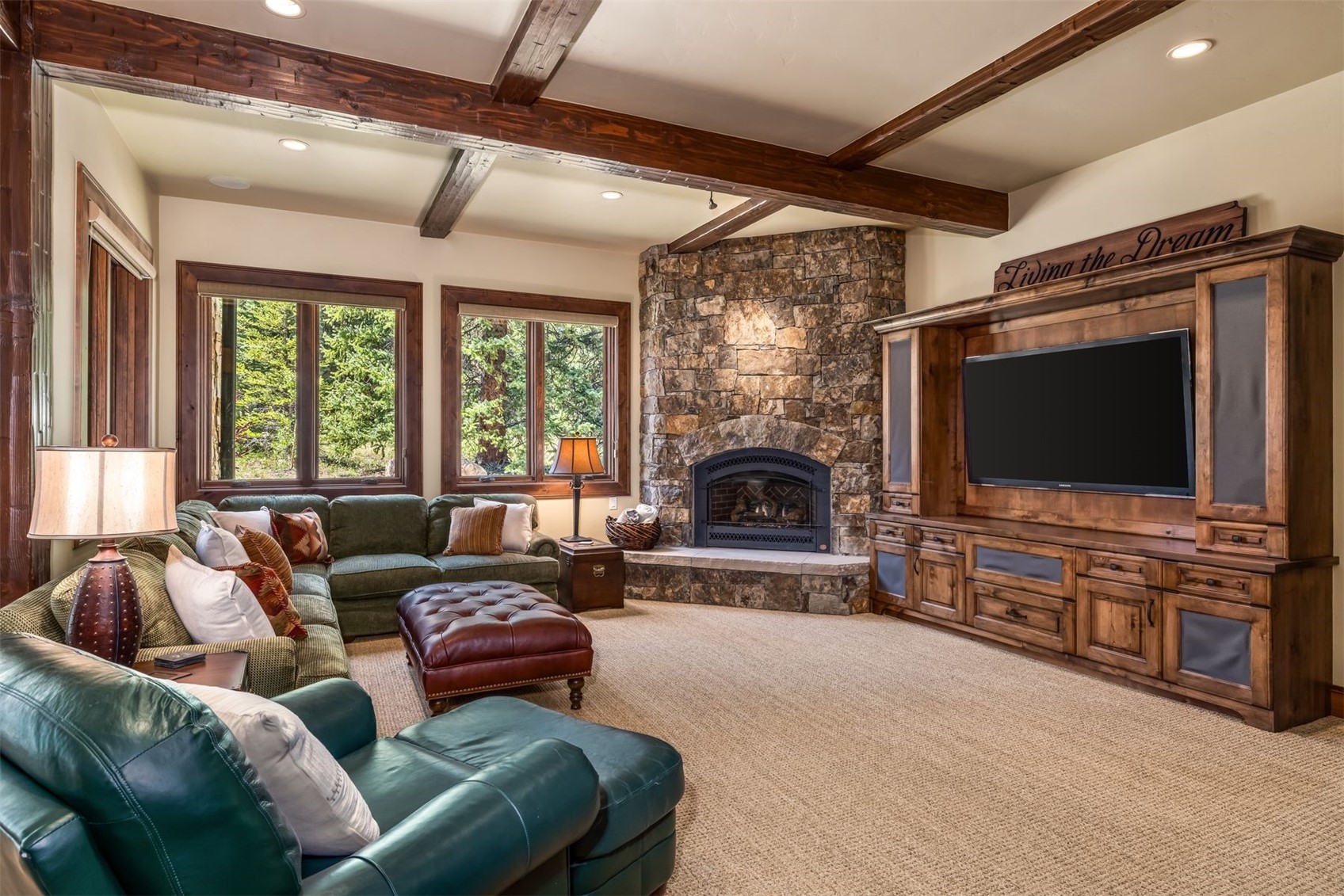 This cozy living room features a rustic aesthetic with exposed wooden ceiling beams and a prominent stone-clad chimney breast housing a fireplace. The space is furnished with a green sectional sofa, a leather ottoman, and a dark leather armchair, all arranged around a large entertainment center. Large windows provide ample natural light and views of the surrounding trees, creating a warm and inviting atmosphere.