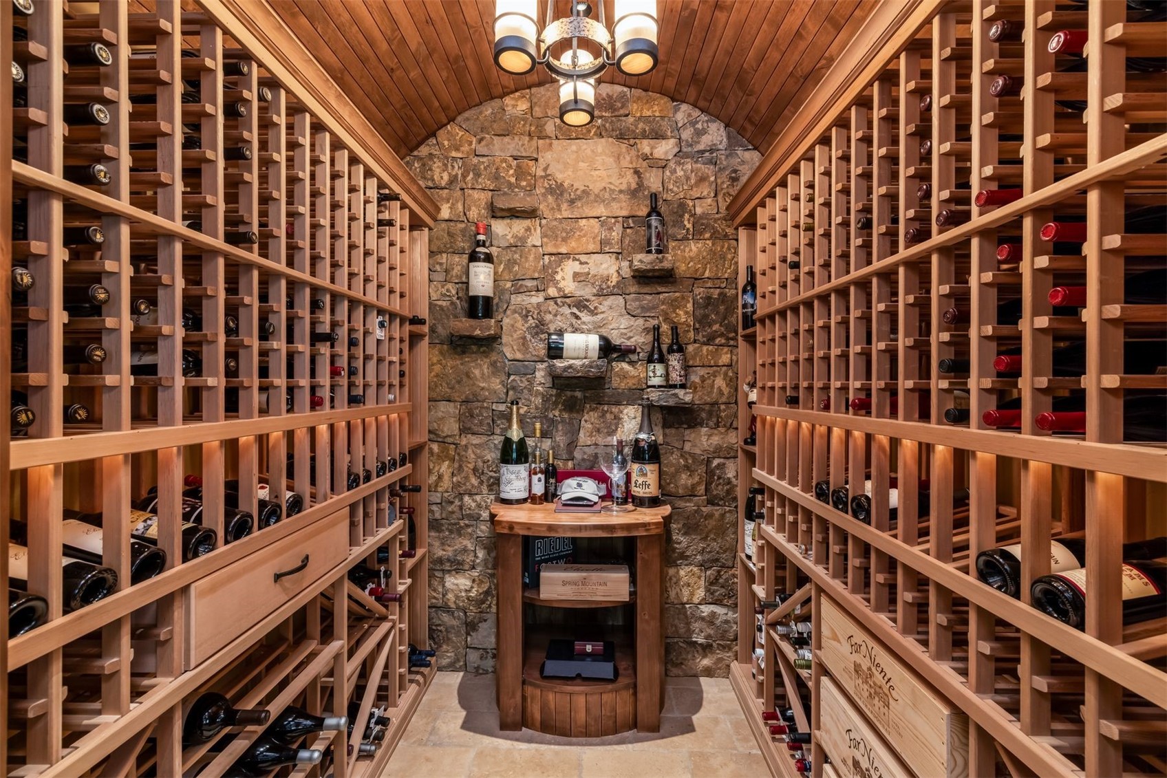 This sophisticated wine cellar features extensive custom wood racking lining both sides of a narrow, arched corridor. The back wall is constructed of rustic, textured stone, providing a dramatic backdrop for a small wooden tasting table and select bottle displays. A decorative chandelier hangs from the barrel-vaulted wood ceiling, creating an intimate and refined atmosphere for wine storage and appreciation.