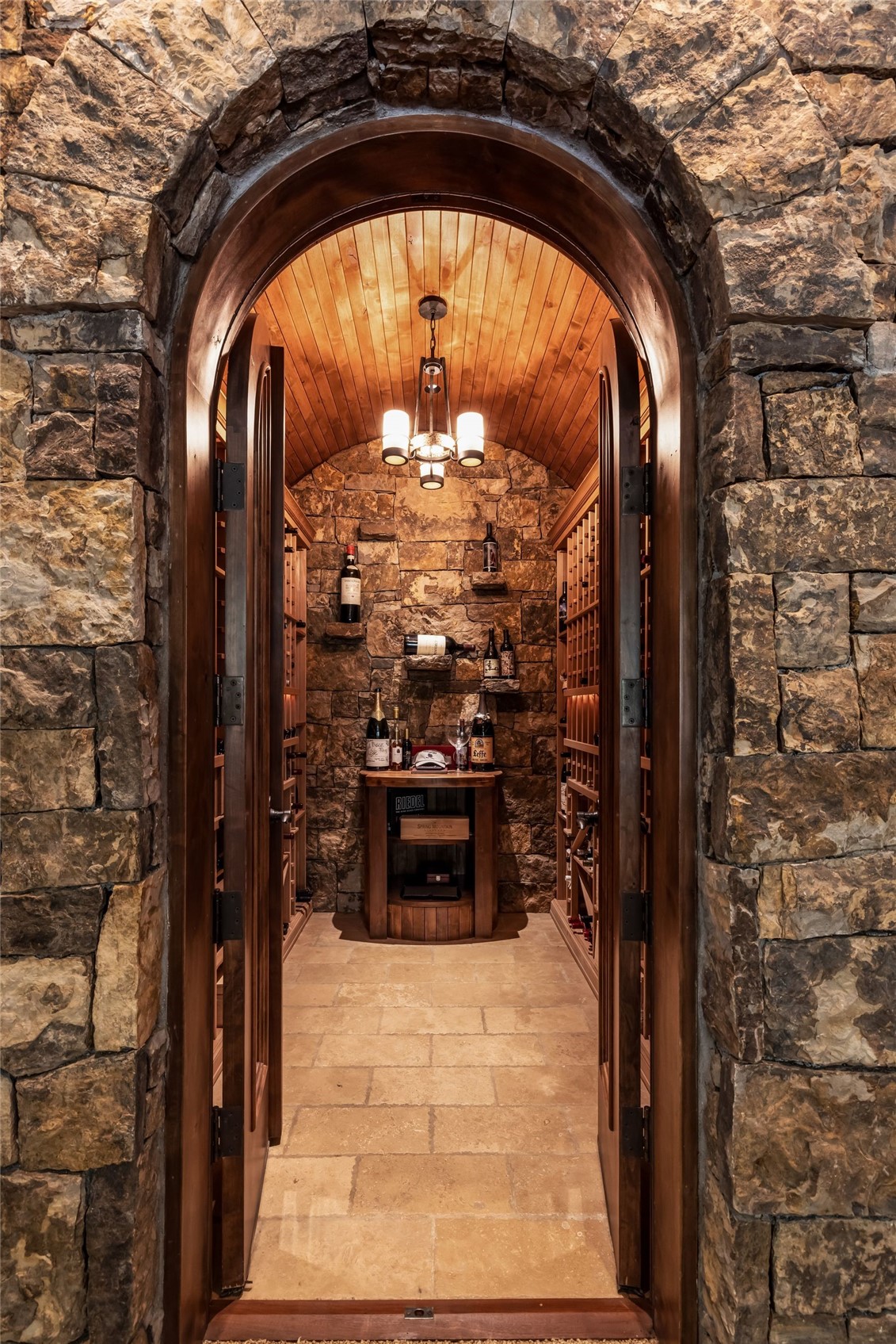 This sophisticated wine cellar features a rustic aesthetic with rough-hewn stone walls and a barrel-vaulted wood ceiling. The space is accessed through a grand arched stone doorway, leading to custom wooden wine racks and a central display table. A classic chandelier hangs from the center, casting a warm, inviting glow over the stone floor and curated bottle collection.