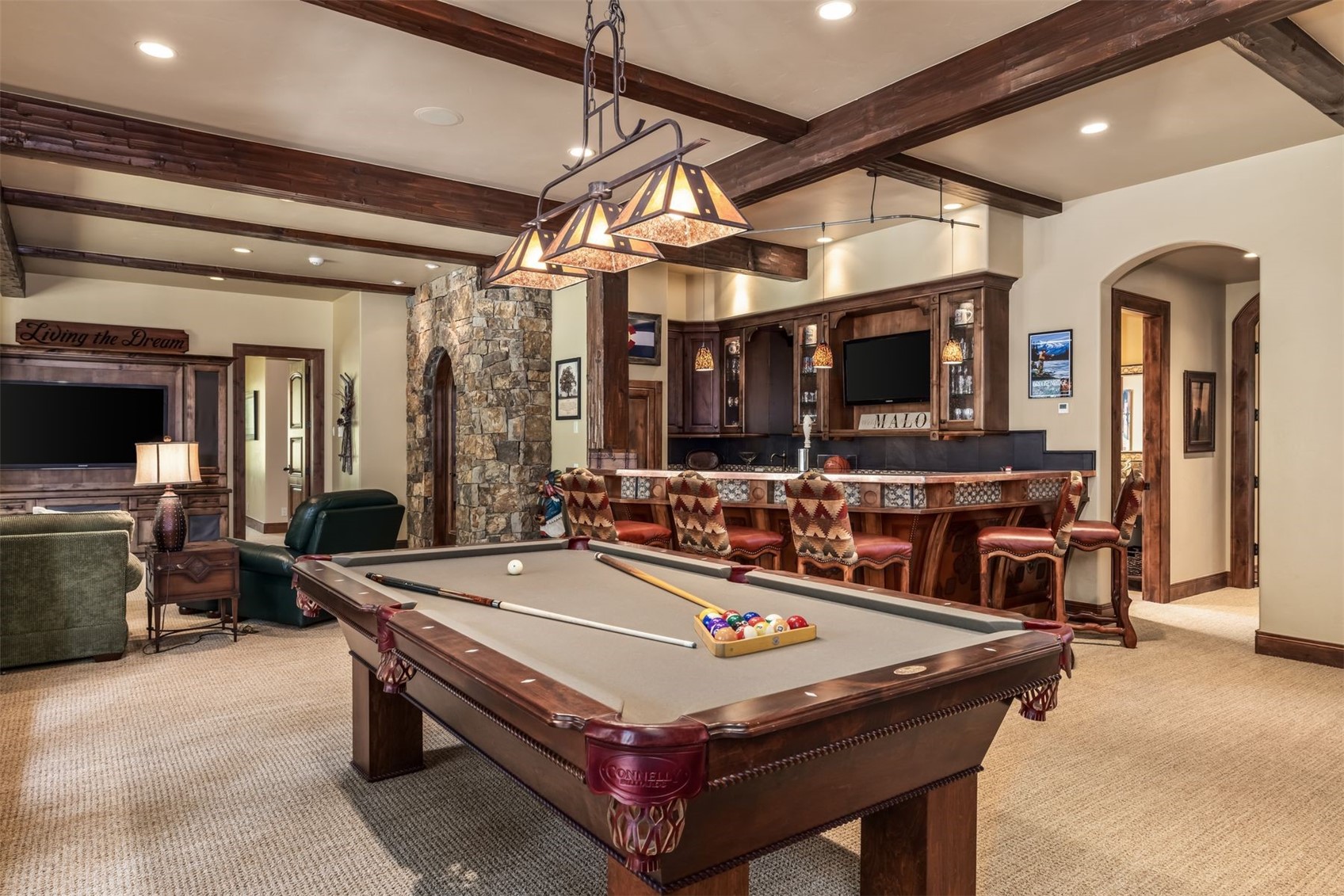 This spacious basement recreation area features a prominent pool table as the central focal point, set against a backdrop of a custom wet bar and a cozy living area. The room is defined by rustic elements, including exposed wooden ceiling beams, a stone-clad wall feature, and warm, patterned upholstery on the bar stools. The overall aesthetic is that of a high-end, lodge-style entertainment space designed for hosting and relaxation.